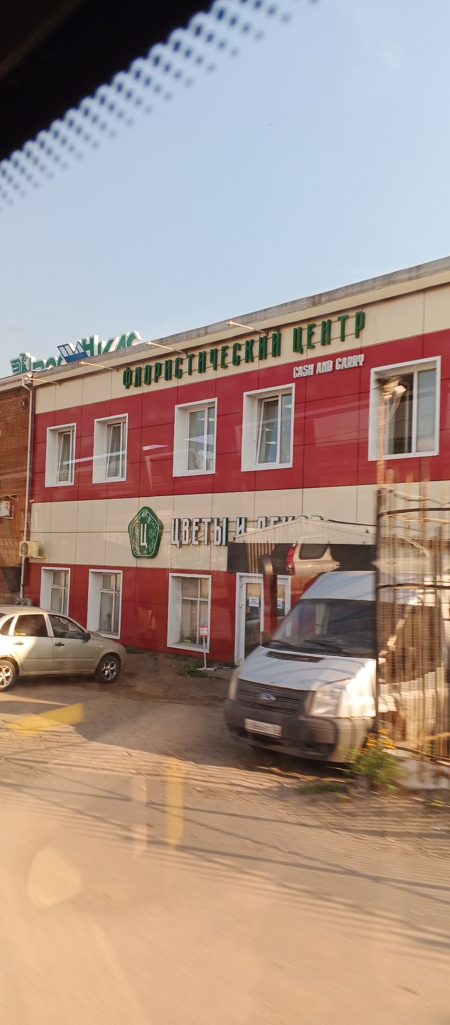 A two-story building with a red and white facade is visible in Izhevsk, Russia. The upper story has green lettering that reads "ФЛОРИСТИЧЕСКИЙ ЦЕНТР" (Floristic Center). Below this, smaller green text says "CASH AND CARRY". On the lower part of the building, there is a green hexagonal logo with a stylized "Ц" inside, next to the words "ЦВЕТЫ И ПОДАРКИ" (Flowers and Gifts). Several windows with white frames are present on both stories.
In front of the building, a silver sedan is parked on the left, and a white van is parked on the right, facing towards the viewer. The van has a license plate that is partially visible. The ground in front of the building is a dirt or gravel surface. The scene is captured from a slightly elevated perspective, possibly from inside a vehicle due to reflections on the windshield. The sky is clear and blue, suggesting daytime.