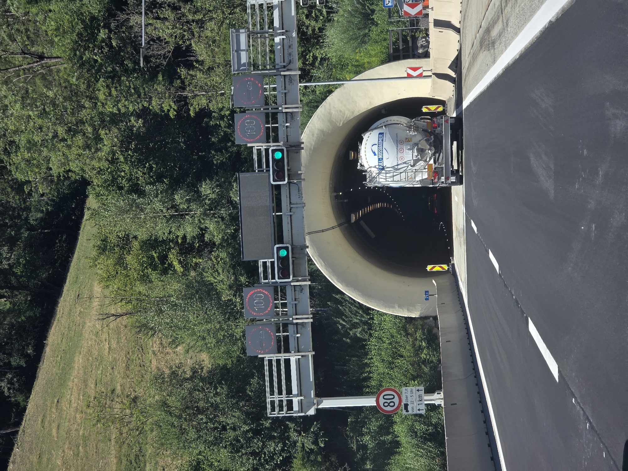 The image shows a forward view of a highway leading towards a tunnel entrance. A white tanker truck with "POLL-NUSSBAUMER" written on its side is entering the tunnel. Above the tunnel entrance, digital signs display "100" and "100" in red, indicating a speed limit. A traffic light with a green signal is visible. To the left of the road, a sign with "80" in a red circle is posted, with additional text below that is partially obscured by the angle but appears to indicate restrictions for vehicles over 7.5 tons between 22:00 and 05:00, with an arrow pointing upwards and "1 km" written. The surrounding environment consists of a grassy hill on the left and dense green trees on the right, suggesting a rural or mountainous area. The road surface is asphalt with white lane markings. The image is taken from a vehicle on the road. This scene is located in Neulassing, Austria, specifically on the A9 Pyhrn Autobahn approaching the Gleinalm tunnel.