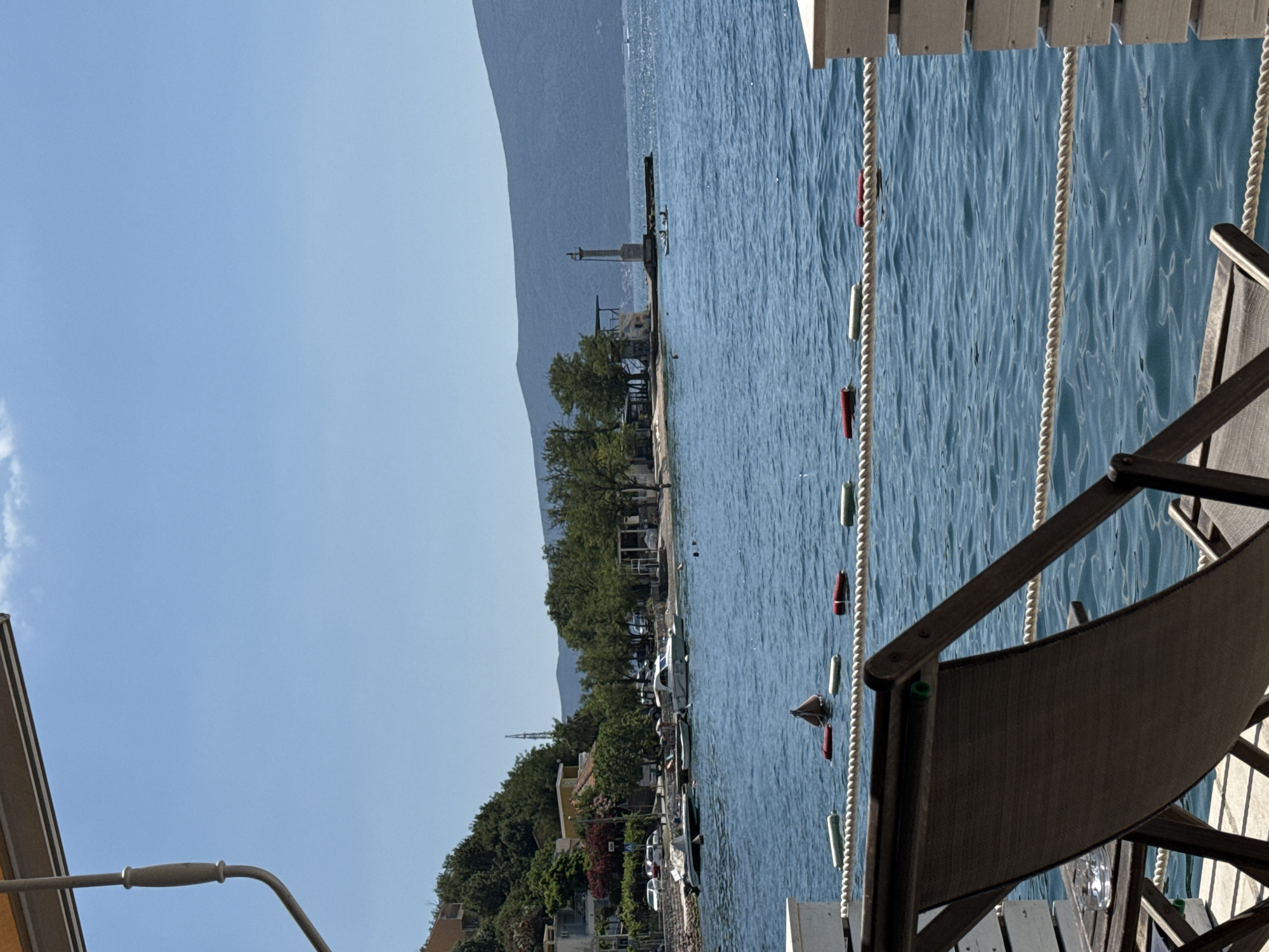 A view of a bay on a sunny day, with a wooden deck in the foreground. On the deck, a lounge chair and a small table with an ashtray are visible. A rope barrier separates the deck from the water. The water is blue and calm, with several buoys and a swimming area marked by a rope. In the distance, the shore is lined with trees and buildings, and a lighthouse stands on a pier. Hills and mountains are visible in the background. The sky is clear and blue with a few wispy clouds.