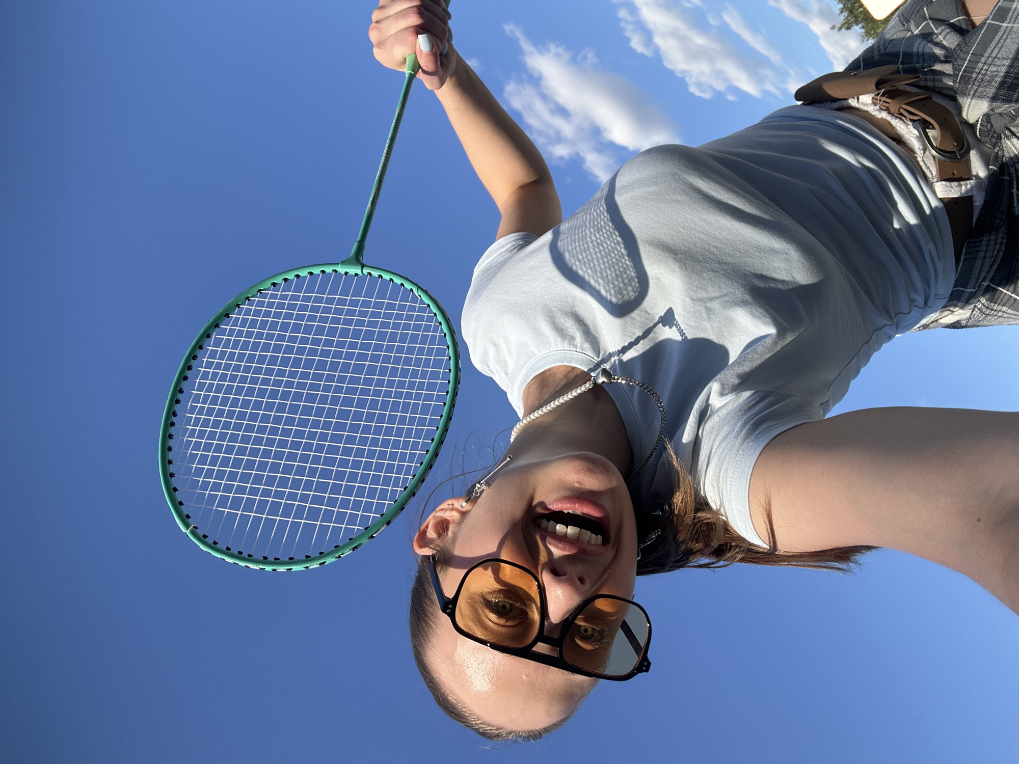 A young woman is holding a badminton racket against a bright blue sky with scattered clouds, suggesting an outdoor setting. She is wearing large, orange-tinted sunglasses, a light blue t-shirt, a pearl necklace, and a plaid skirt with a brown belt. Her right arm is raised, holding the racket by its handle. Her expression is one of joy, with her mouth open and teeth visible. The image appears to be a selfie taken from a low angle, emphasizing the sky and the woman's playful pose. The location is specified as the city of Khimki, Russia.