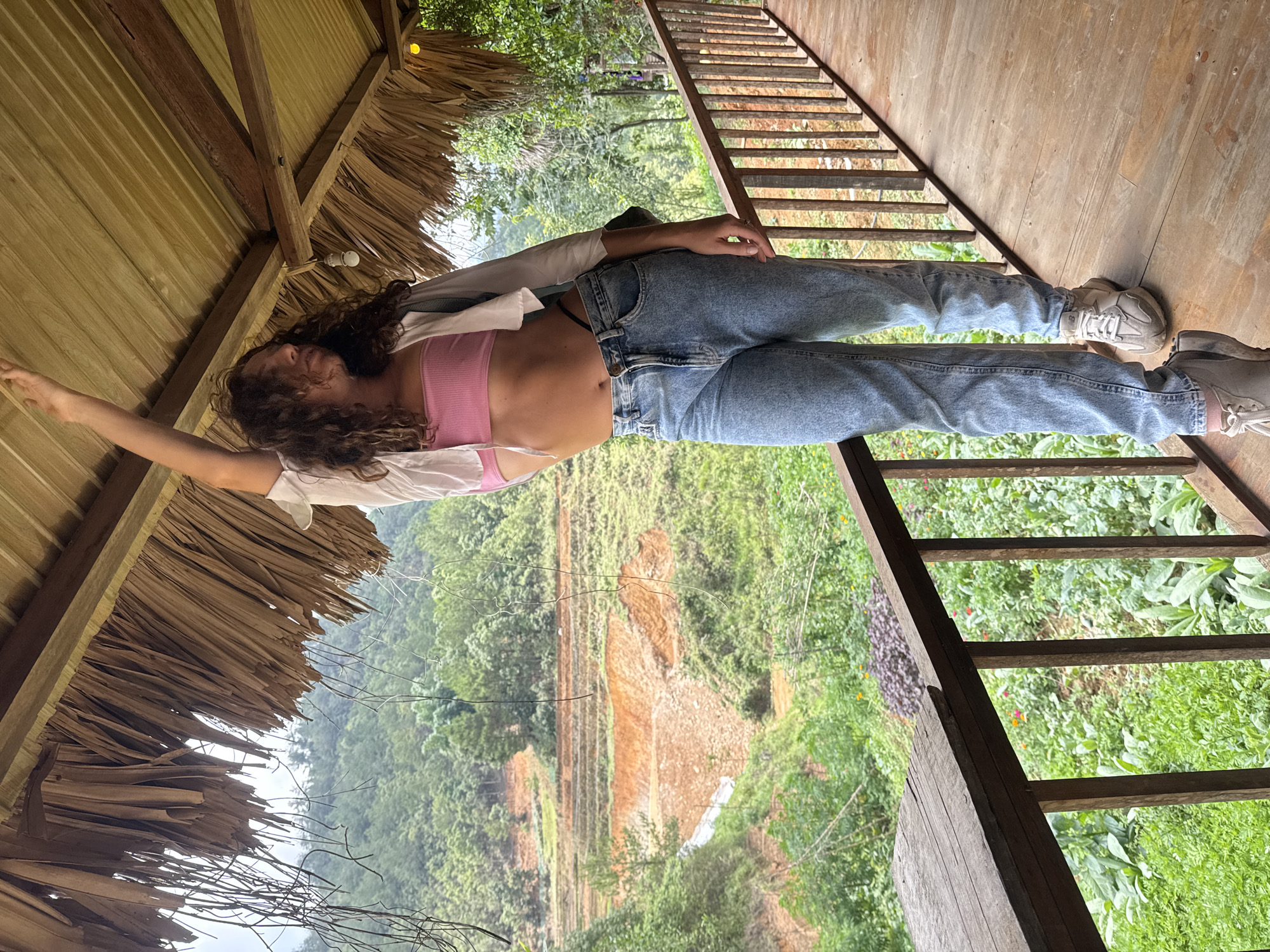 A woman is lying on a wooden platform with a view of a hillside near Ngã Ba Kim, Vietnam. The woman, positioned at an angle in the frame, has curly brown hair and is wearing a pink crop top, a white shirt, and blue jeans. Her left arm is extended upward towards the thatched roof of what appears to be a hut or shelter. Her feet are towards the right side of the image, and she is wearing grey sneakers.
The platform is constructed of wood and features a railing made of horizontal wooden bars. The background showcases a lush, green hillside with what appears to be terraced or altered land in a reddish-brown color. The environment is indicative of a tropical or subtropical climate. A portion of the roof of the shelter is visible above the woman’s head and to the left of the frame.