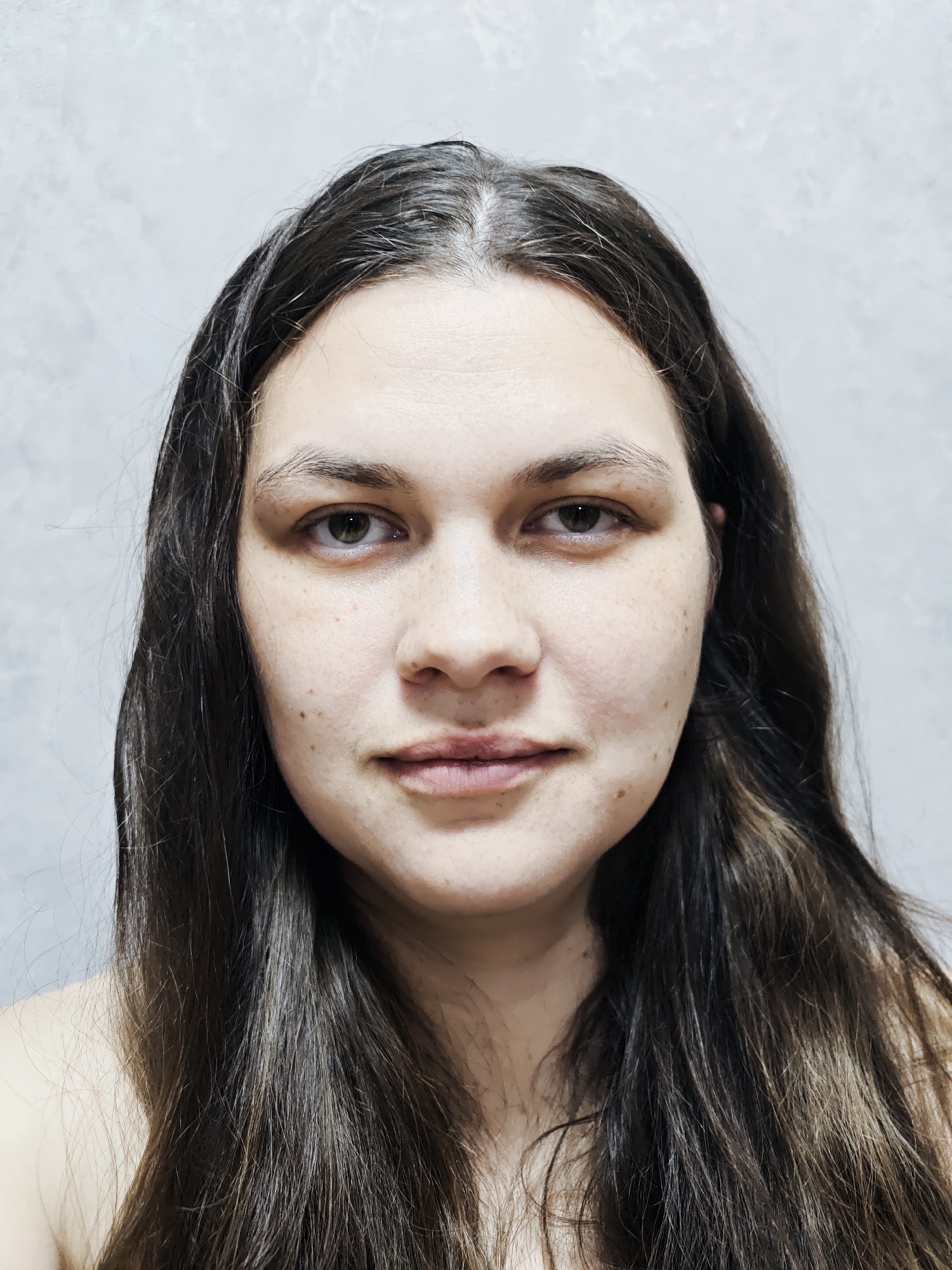 A close-up portrait features a young adult woman looking directly at the camera. She has long, dark brown hair, which appears slightly disheveled and has noticeable grey strands, particularly at the roots along her central parting. Her skin is fair with numerous small freckles and a few moles visible across her face, suggesting a natural, unadorned appearance. Her dark brown eyes are open and gaze straight ahead. Her expression is neutral, with a very subtle, almost imperceptible upturn at the corners of her lips. There are faint lines on her forehead and slight shadows under her eyes. Her nose is straight, and her lips are naturally full and slightly parted. The woman's bare shoulders are visible at the bottom of the frame, suggesting she might be wearing a strapless garment or her shoulders are uncovered. The background is a light grey, textured wall, providing a simple and unobtrusive setting. The lighting is even and soft, illuminating her face without harsh shadows. No specific activity is taking place other than posing for a portrait. There is no visible text in the image.