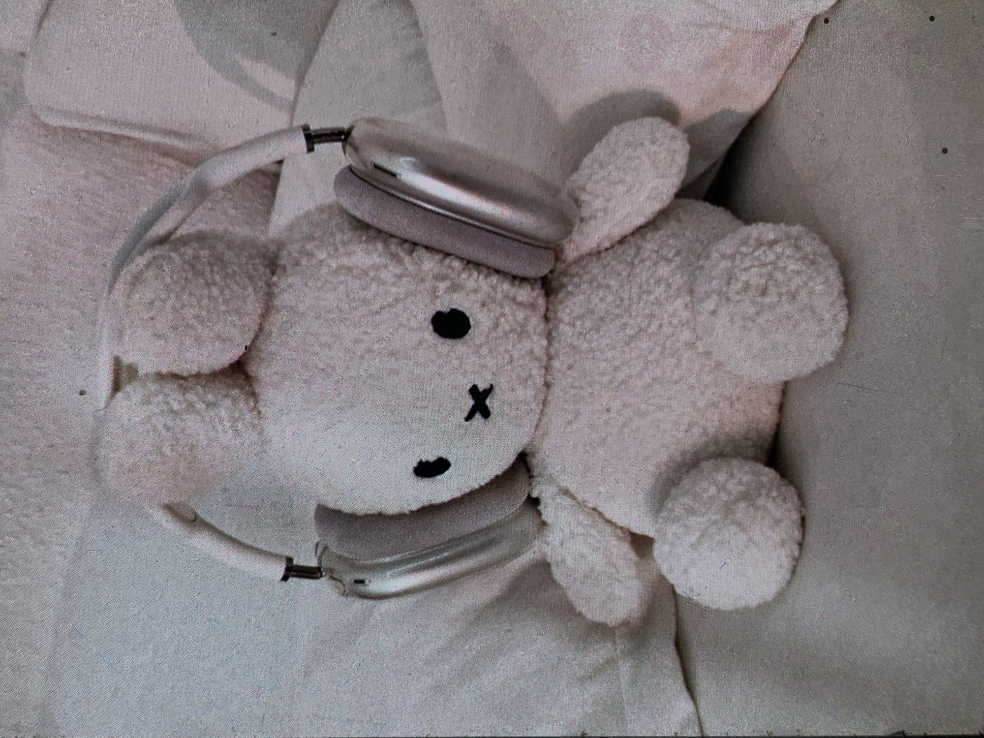 A stuffed bunny toy wearing silver over-ear headphones is positioned centrally on a light-colored, textured surface, likely a bed or couch. The bunny is fluffy and white, with two small, rounded ears, black button eyes, and a small black "x" for a mouth. The headphones appear to be a consumer electronic device, with silver earcups and a white headband. The background consists of soft, crumpled fabric in a similar light color. There is no visible indication of the city of Nizhny Tagil, Russia, or any elements that would place the scene in that specific geographical location. The image is somewhat grainy and has a soft, diffused lighting.