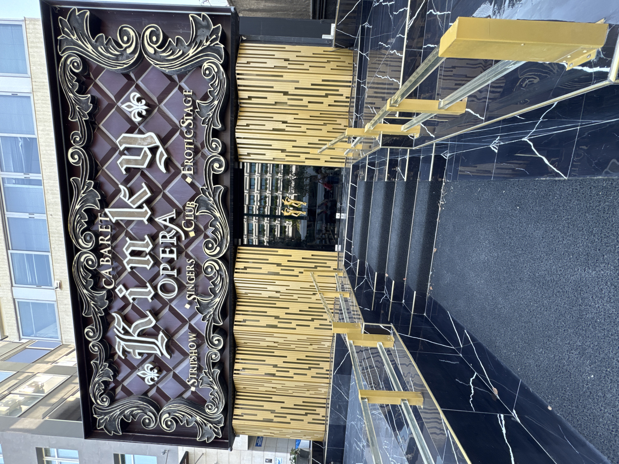 A wide shot captures the exterior entrance of a club called "Kinky Opera" in Athens, Greece. The building's facade features a prominent sign with the club's name in large, ornate lettering, surrounded by decorative scrollwork. Below the name, smaller text indicates "CABARET," "STRIPSHOW," "SINGERS," "CLUB," and "EROTIC STAGE." The entrance itself is set behind a wall of vertical golden slats, with a glass door. A set of black marble stairs with a black carpet runner leads up to the entrance, bordered by a railing with gold-colored posts and clear glass panels. The marble flooring has a distinctive white veining pattern. The overall scene suggests a nightlife venue with a visually striking and perhaps opulent aesthetic.