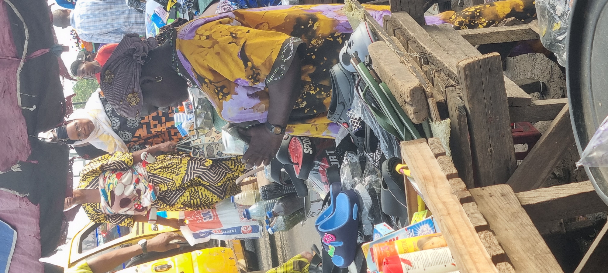 The image depicts a bustling commercial scene in Lagos, Nigeria. A woman wearing a yellow and purple patterned garment and a headwrap is the central figure. She appears to be engaged in commerce. To her right, there is a collection of merchandise including footwear and household items arranged on a wooden structure. Additional individuals are visible in the background, suggesting a crowded or marketplace environment. A yellow vehicle is seen in the lower left corner, further suggesting an active urban setting.