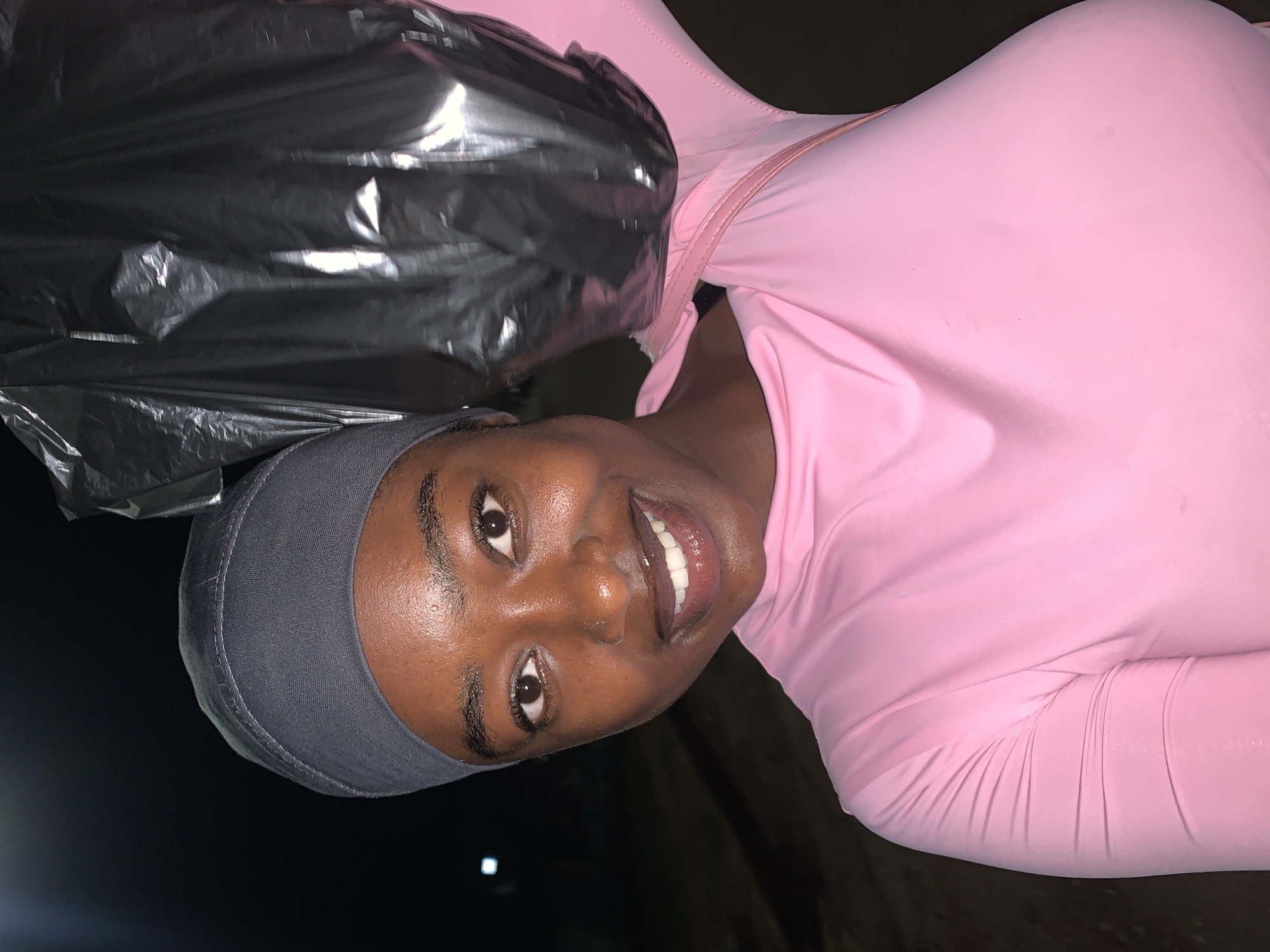 A woman is smiling and looking at the camera. She is wearing a gray cap, a pink long-sleeved shirt, and has a silver bag draped over her shoulder. The background is dark and indistinct. The location is potentially the city of New Stadium, Nigeria, but there is no definitive information in the image to confirm this.