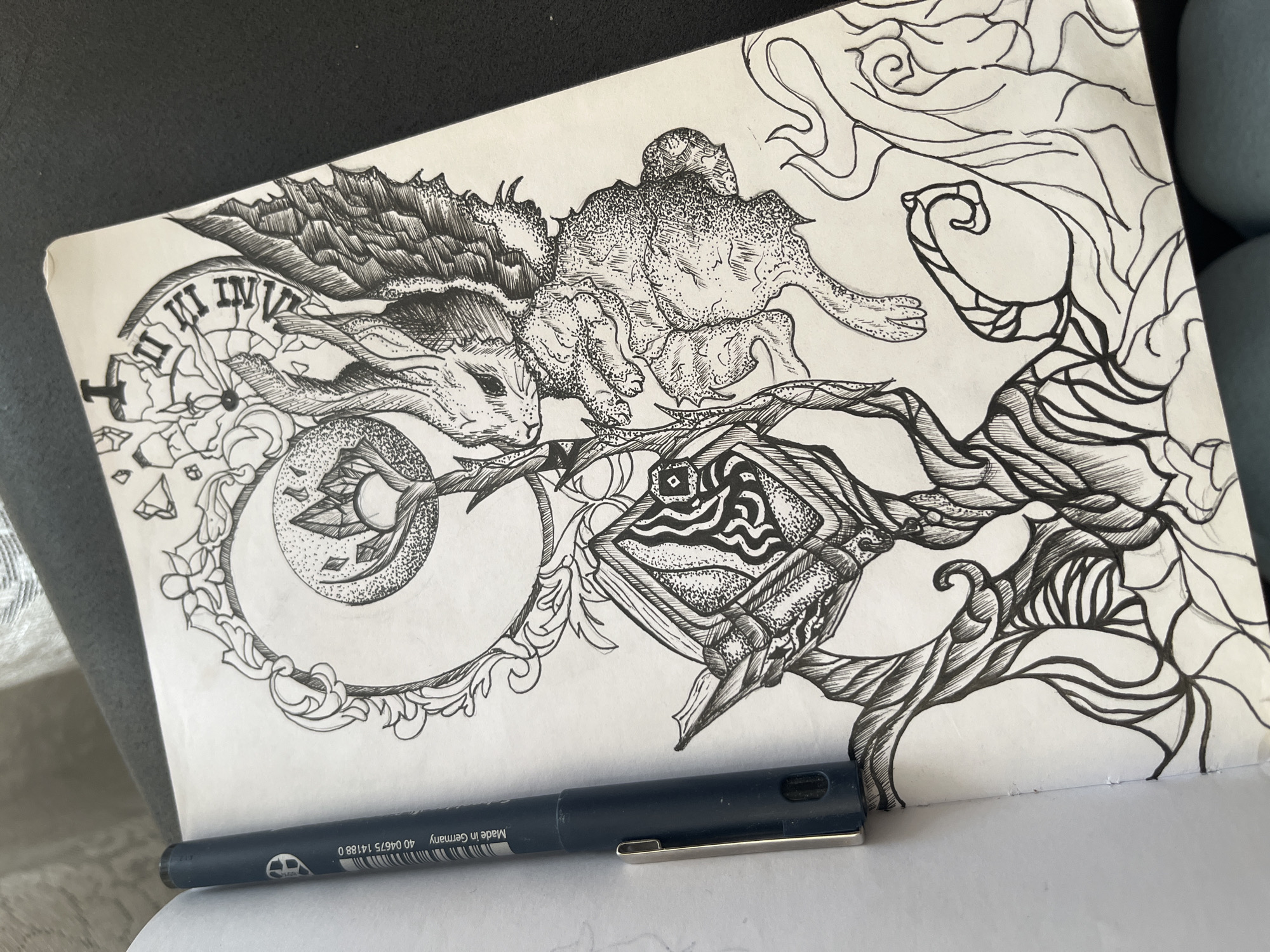 The image shows a black and white ink drawing on a sheet of paper. The drawing features surrealistic elements, including a rabbit-like creature with wings positioned next to a quadruped with a spiky back, possibly a reptile. Below the rabbit, there's a broken clock face with Roman numerals, and to the right of the creatures, there's an object resembling a stylized fish head surrounded by winding, tree-like branches. A black pen with the inscription "Made in Germany" lies at the bottom edge of the paper. The background is out of focus, showing a couch or other upholstered furniture. There are no clear indications of Sofia, Bulgaria.