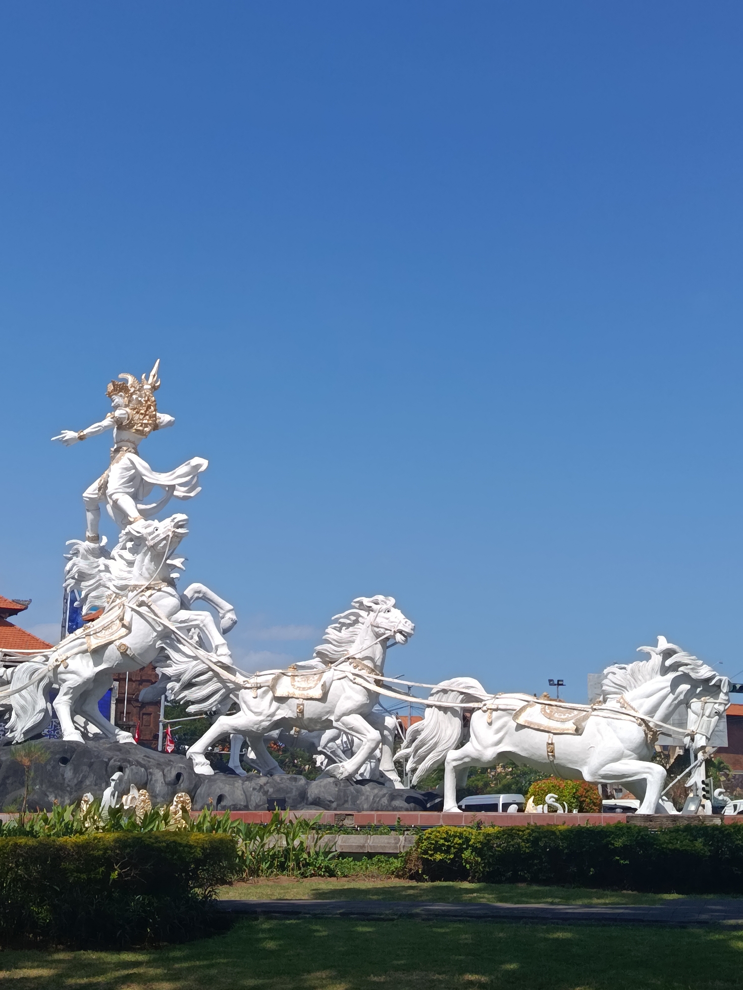 The image shows a large white statue of a deity riding a chariot pulled by multiple horses. The deity is adorned with golden headwear and jewelry and is in a dynamic pose, with one leg extended. The horses are also white, with flowing manes and tails, and are harnessed together to the chariot. The statue is situated in a roundabout in Kuta, Indonesia, with a clear blue sky as the background. In the foreground, there is green grass and some bushes. The statue is a prominent landmark and a popular tourist attraction in the city.