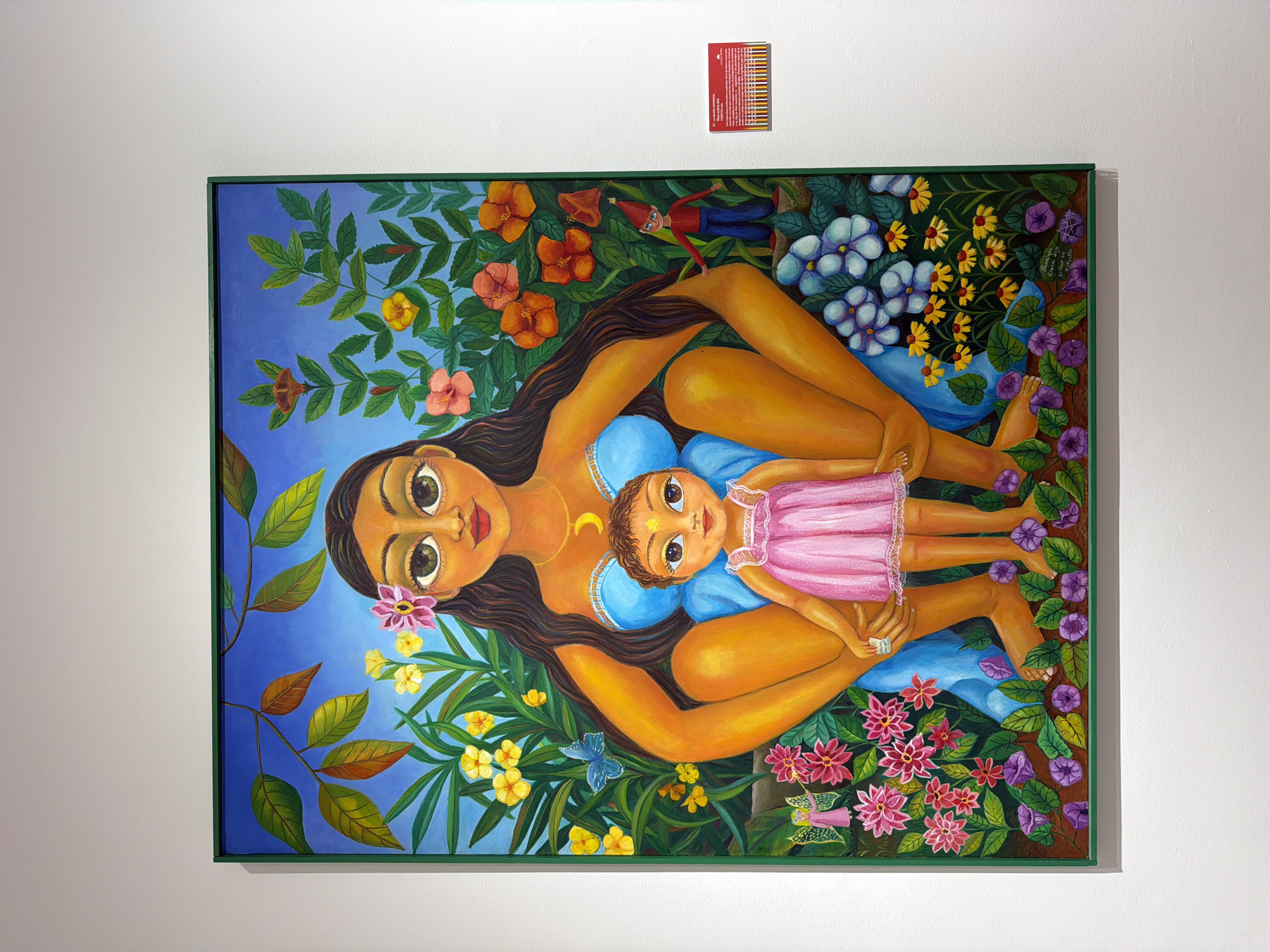 A framed painting hangs on a white wall in what appears to be a gallery. The painting is of a woman and a child surrounded by vibrant flowers and lush greenery. The woman is depicted in a squatting position, embracing the child who is wearing a pink dress. Both figures have large, expressive eyes. The painting's frame is a bright green. To the right of the painting, a small red plaque with text is attached to the wall. The text on the plaque is illegible in the image. The scene is lit by artificial light, casting a soft glow on the artwork and wall.