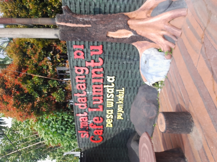 The image shows a sign in front of a cafe in Kecamatan Pujon, Indonesia (coordinates: -7.855, 112.455). The sign reads "slmat datang di Cafe Lumintu desa wisata pujon kidul" in red and yellow lettering on a dark green brick wall. The sign is partially obscured by a large tree trunk, which appears to be made of wood and painted brown. The tree trunk is positioned to the right of the sign and extends to the ground, with visible roots. In front of the sign are two round, flat, wooden tables, and a small wooden stump. There are several large rocks in the background. The ground is covered in wooden planks. There are no people visible in the image. The environment is lush, with green foliage and tree branches visible in the upper portion of the image. The image appears to have been taken during the day.