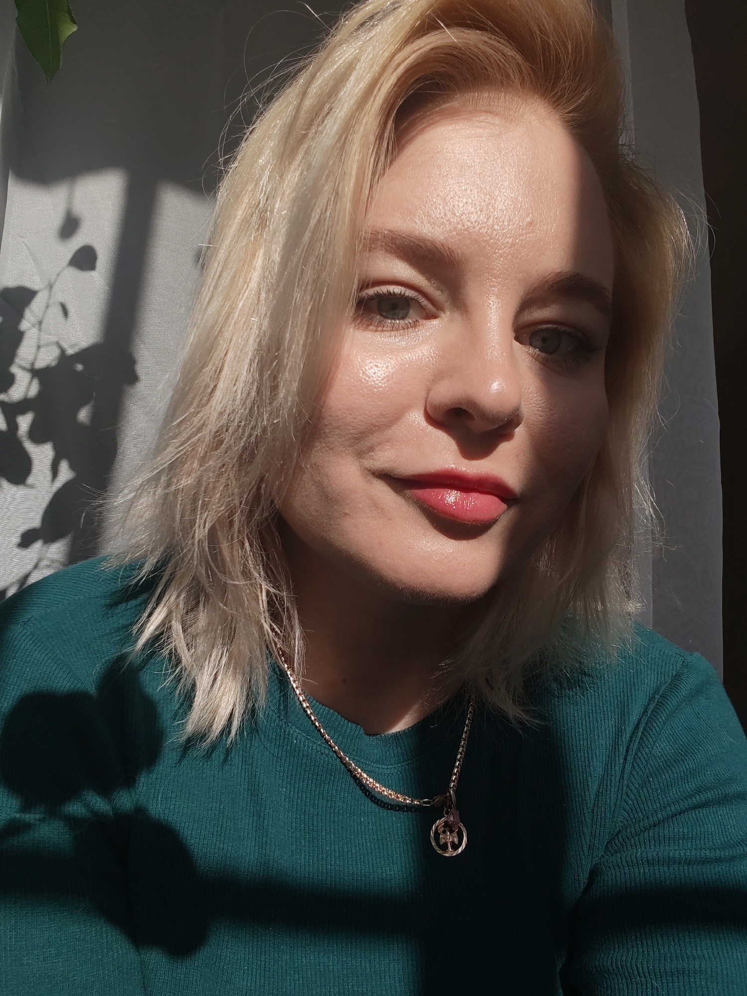 A close-up portrait shows a woman with blonde hair and green eyes, wearing a green top and a gold necklace with a round pendant. She is smiling slightly, with red lipstick. The lighting is dramatic, with strong shadows on her face and clothing. The background includes a white curtain and the shadow of a plant. There is no information to connect this image to Izhevsk, Russia.