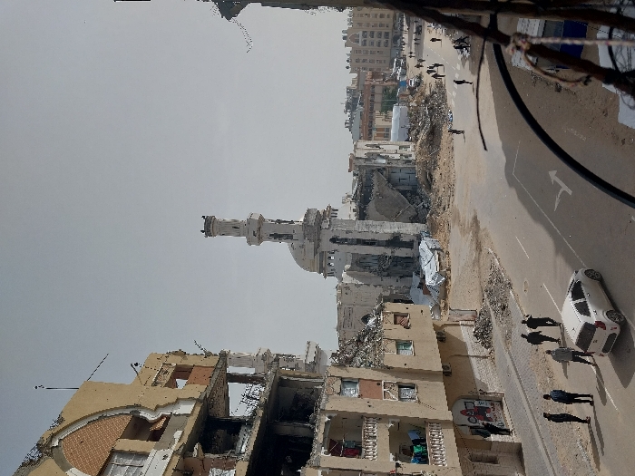 This elevated view captures a scene of widespread urban destruction in Khan Yunis, Palestinian Territory. In the center-background stands a severely damaged mosque, its dome partially collapsed and its minaret showing significant wear, surrounded by large piles of rubble and debris. To the immediate left, a multi-story building, likely residential, exhibits extensive structural damage with large sections of its facade missing, revealing exposed interiors, rebar, and balconies, some with hanging laundry. The ground floor of this building features a storefront with visible graffiti.
An asphalt road runs across the mid-ground, featuring white right-turn arrows. On the left side of the road, a white compact car, possibly a Kia Picanto, is present, and a group of approximately five men stand on the adjacent sidewalk. Further into the background, several more individuals and small groups, totaling around 15-20 pedestrians, are visible walking on streets amidst other damaged buildings and scattered debris. The sky is overcast and light grey. Partially visible in the lower right foreground are out-of-focus dark cables or structural elements, indicating the vantage point.