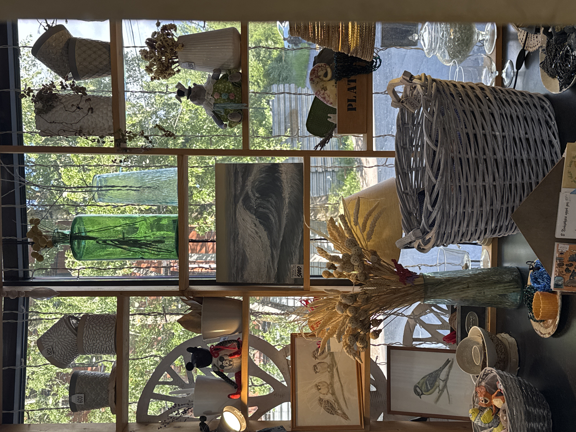 A sunlit indoor scene displays a collection of decorative items arranged on wooden shelves in front of a window. The window overlooks a leafy green outdoor environment with trees and a fence. The items on display include several ceramic pots, a green glass vase with dried reeds, a light blue glass vase, a white textured vase with a trailing plant, a stack of patterned pots, a white vase with dried flowers, a stuffed toy resembling a hippopotamus wearing a hat and clown-like costume, and a framed painting of a wave. On lower shelves are a framed drawing of sparrows, another framed drawing of a bird, a basket containing small toys and decorative items, a large woven gray basket, a wooden crate with the word "PLAT" visible, a glass jar with a lid filled with small green spheres, and various other decorative objects and bowls. The overall impression is of a shop or display area filled with rustic and handcrafted goods.