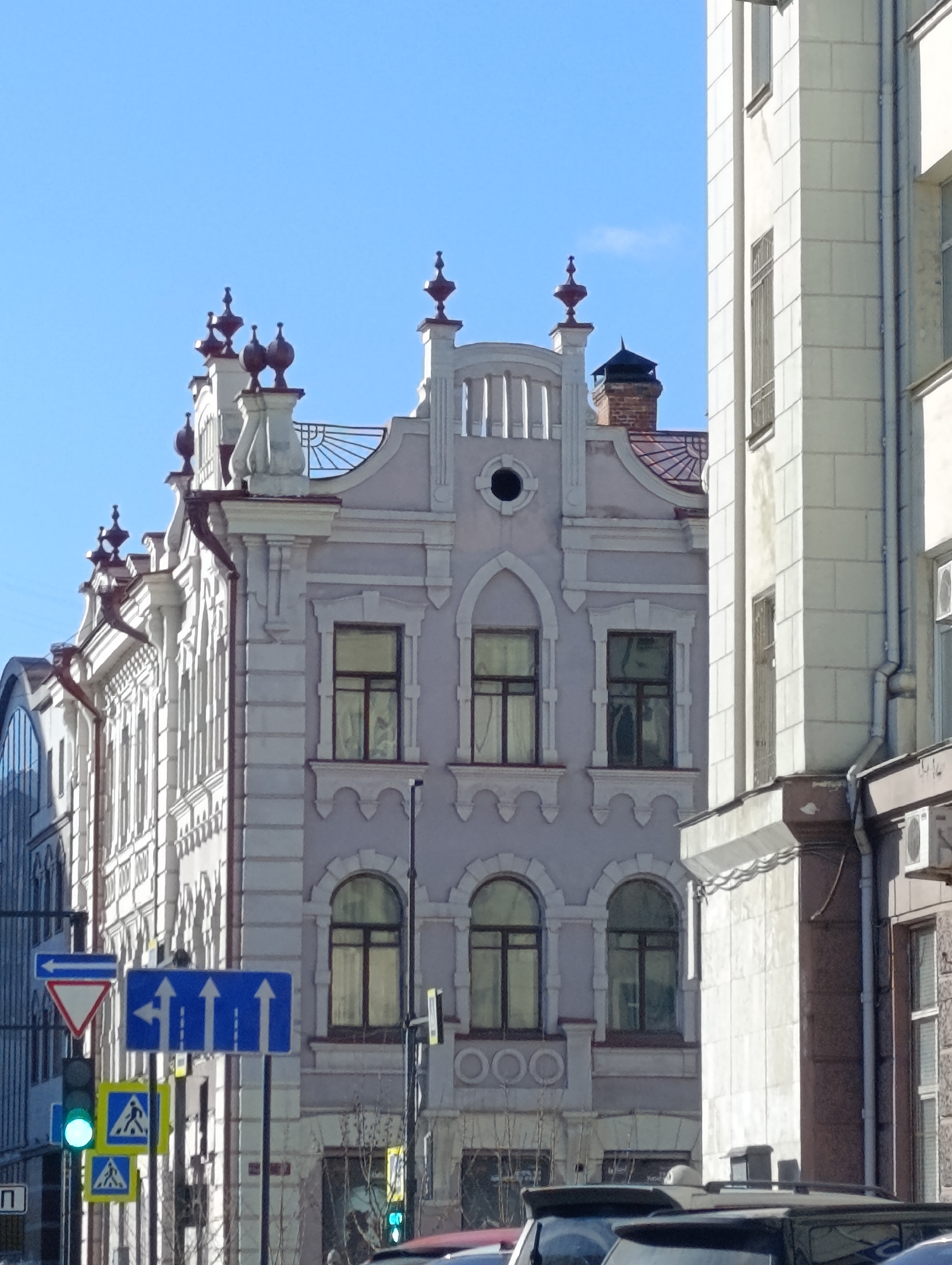 The image presents a street view in Krasnoyarsk, Russia, focusing on two prominent buildings against a clear blue sky. The building in the center is a three-story structure with ornate architectural details, including decorative spires, arched windows, and a distinctive parapet. The building is painted in a light color. To the right, a taller, white building with a more modern design partially obscures the central structure. Various street signs are visible, including a traffic light showing a green pedestrian crossing signal and blue signs indicating lane directions. Vehicles are visible at the bottom of the frame, suggesting a busy street. There is no people in the image.