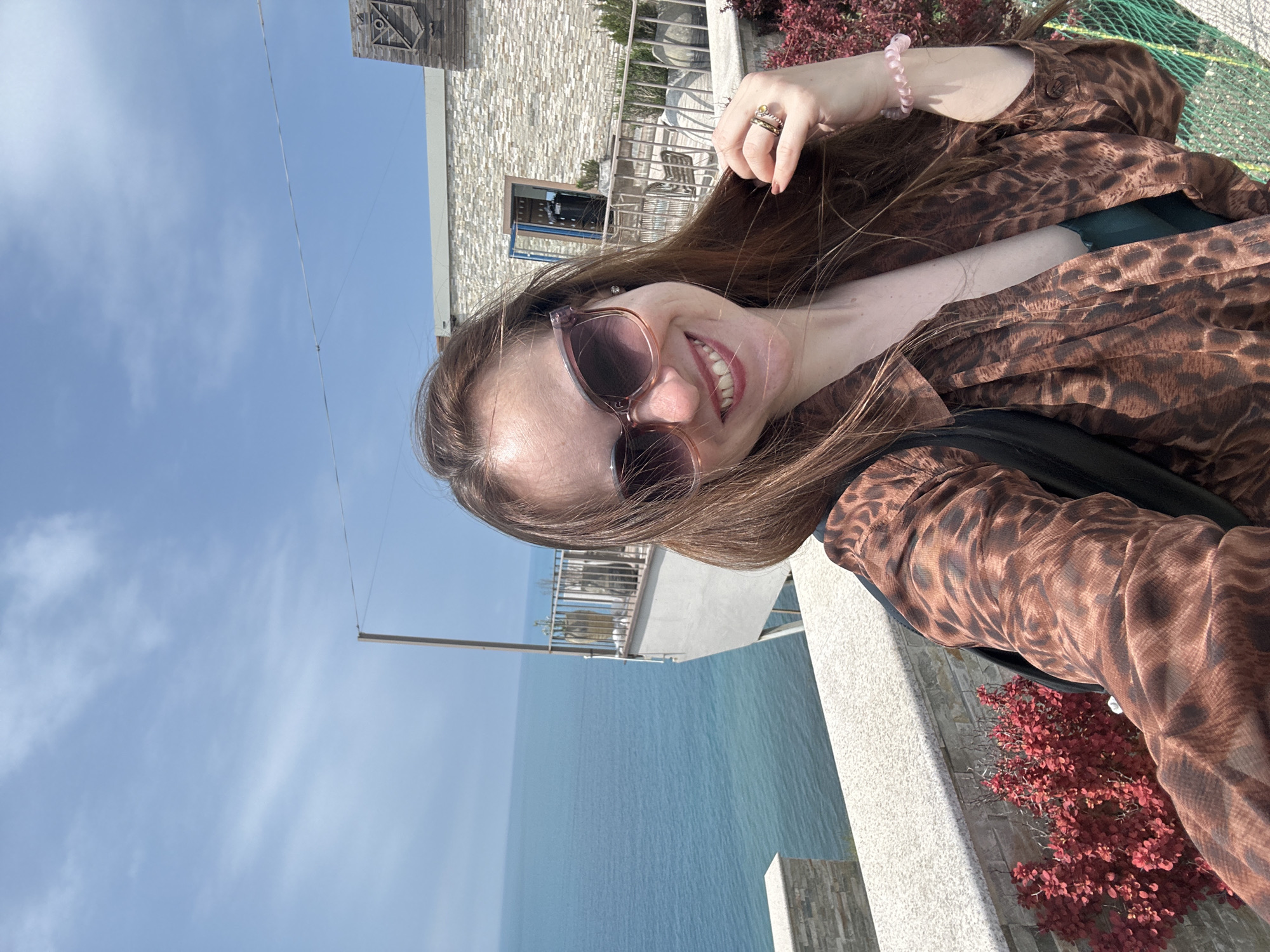A woman is taking a selfie outdoors on a sunny day. She is wearing sunglasses and a brown leopard-print shirt, with a pink bracelet on her wrist and rings on her fingers. Her hair is long and brown. Behind her is a stone building, a metal railing, red bushes, and a body of water, presumably the sea, under a blue sky with some clouds. The location is likely in Anapa, Russia.