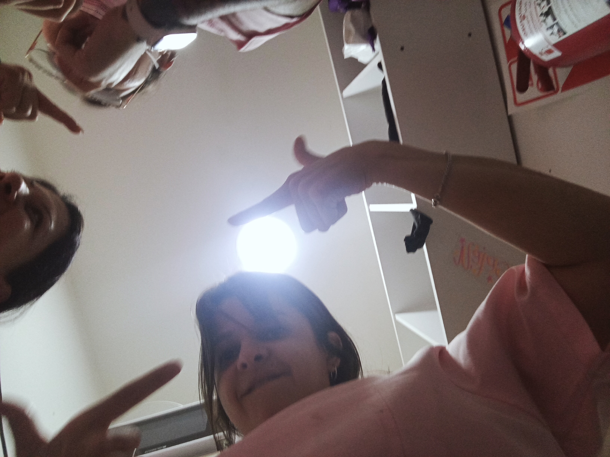 The image presents a low-angle, upward-facing shot of three individuals. Two are partially visible, with one appearing in the lower left and another near the top. The third person occupies the central foreground. All three are pointing towards the camera. The background reveals elements of an indoor setting, including shelves, clothing, and a fire extinguisher. There are no indications of the city of Buenos Aires, Argentina, in the visual information provided.