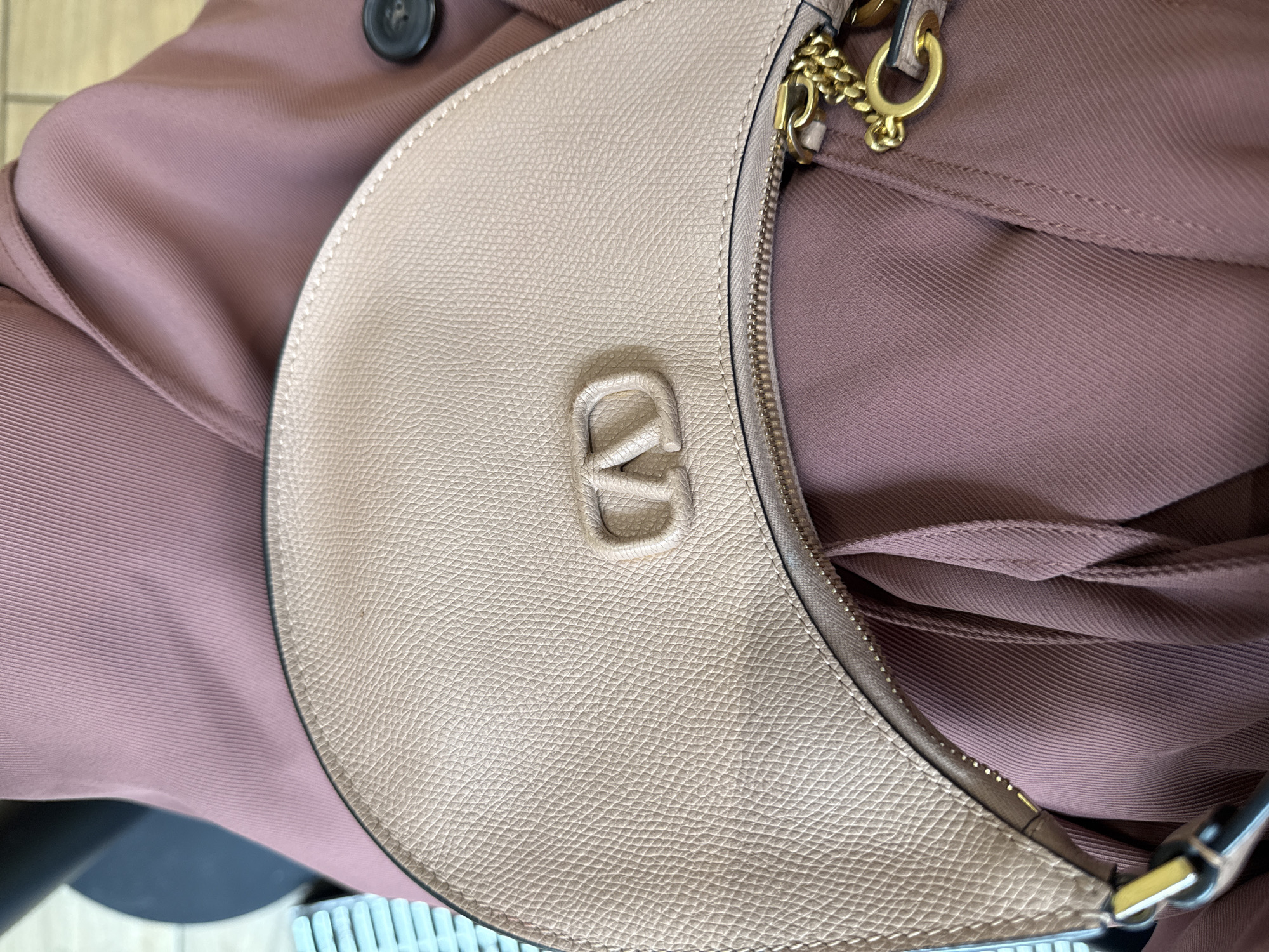 This close-up, top-down photograph presents a light beige or tan handbag with a distinctly textured surface, likely pebbled leather. The handbag features a crescent or half-moon shape. A prominent, embossed 'V' logo is centered on the front, suggesting a specific designer brand. Gold-toned hardware, including a zipper and parts of a chain strap, adds metallic accents to the bag.
The handbag is positioned on a piece of clothing, possibly a jacket or blouse, rendered in a muted mauve or dusty rose hue. This fabric exhibits fine vertical ribbing or subtle pleating. A dark, possibly black, button is partially visible on the fabric in the upper left area of the frame. In the lower background, there are glimpses of a light blue/white ribbed structure, potentially a radiator or window blinds, along with a dark, indistinct object.
The scene appears to be indoors, under soft lighting that emphasizes the tactile qualities of the handbag's texture and the fabric's weave. No individuals are present in the image, nor is any specific activity depicted. The only visible emblem is the 'V' logo. While the location context is provided as Moscow, Russia, the image itself does not contain any explicit geographical markers or external views to confirm this. The overall aesthetic is clean and focuses on materials and subtle color coordination.