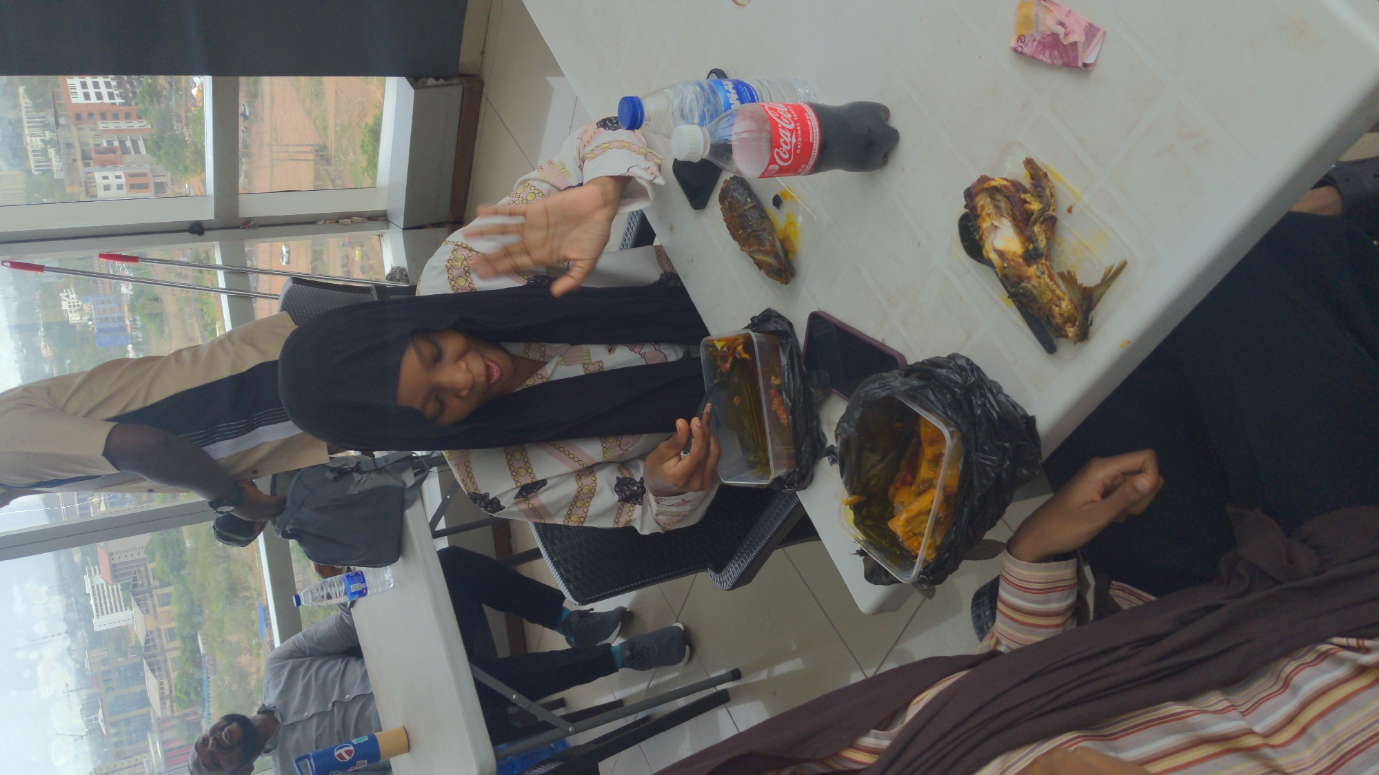 The image captures an indoor gathering of several individuals around a white table, overlooking an urban landscape through large windows. The scene is brightly lit by natural light, suggesting daytime with an overcast or hazy sky.
At the center, a woman in a black hijab and a white and gold patterned top smiles and gestures with an open palm, holding a smartphone in her other hand. To her left, partially visible, a man in a beige and black shirt stands near the window. Further down the table, a man with a beard, wearing a grey shirt, is seated, with a blue and white container in front of him. Another person in a striped top is partially visible in the foreground, with their arm resting on the table.
The white table holds various items, including food and drinks. There are bottles of Coca-Cola and Aquafina water. Two portions of grilled fish are visible; one is in a clear plastic container, while the other is on a black plastic bag. A clear plastic container filled with a greenish-yellow dish, possibly moin-moin, is also on the table, next to a black plastic bag that likely contains more food. A stack of pink currency notes, likely Nigerian Naira, rests near one of the fish portions. Several smartphones are also present on the table. A grey backpack is visible on the floor next to one of the chairs.
In the background, beyond the large windows, a city skyline stretches out, featuring various buildings, some appearing to be under construction, and patches of undeveloped land. Mops or cleaning tools are leaning against the window frame. The overall atmosphere appears relaxed and social, with the individuals engaging in conversation and sharing a meal. The visible text includes "Coca-Cola" on the soda bottle and "Aquafina" on the water bottle. The high vantage point suggests the gathering is taking place in an elevated building within Abuja, Nigeria.