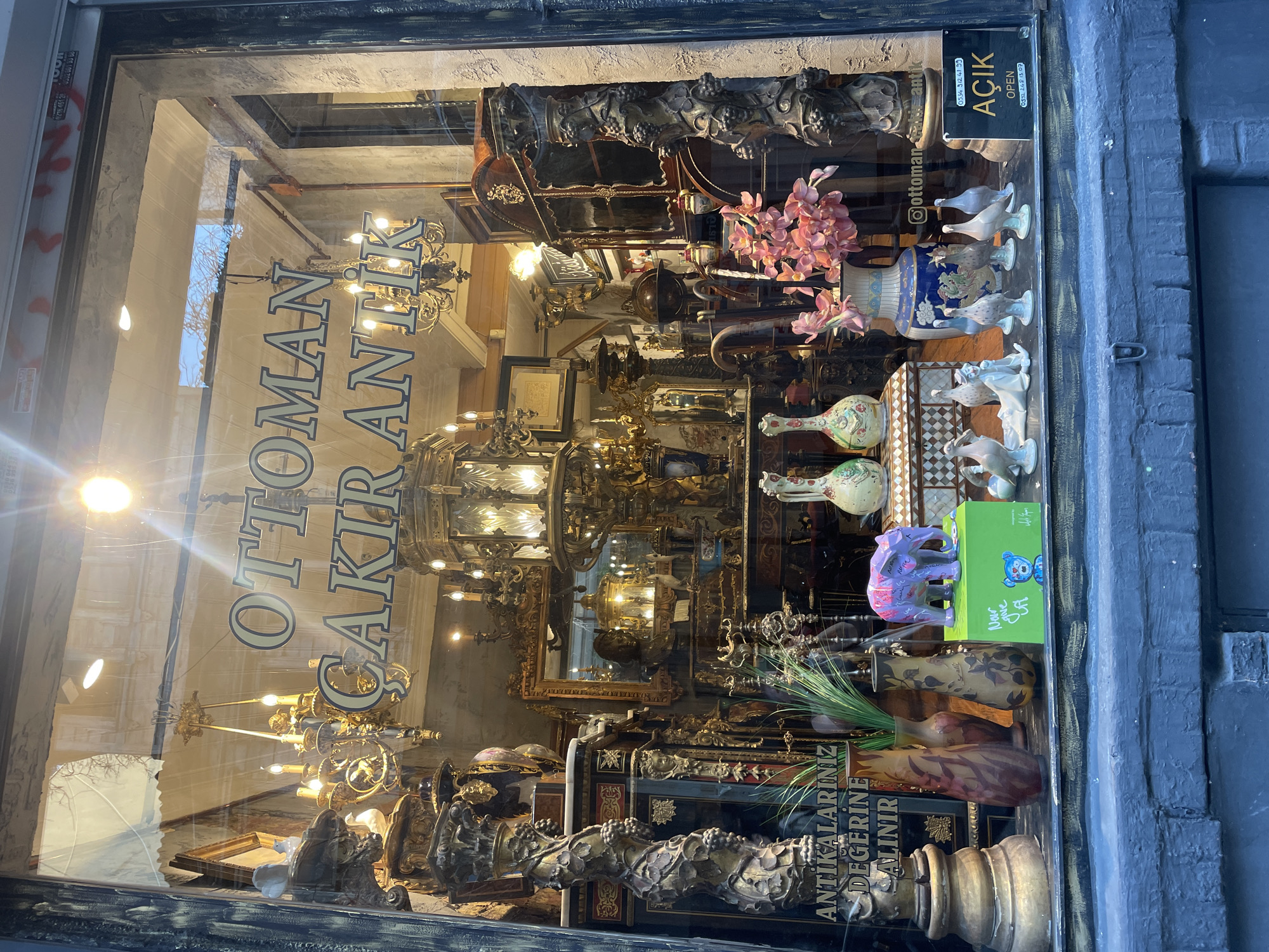 The image shows a storefront window display for an antique shop named "Ottoman Çakır Antik" in Kadıköy, Turkey. The window is filled with a variety of antique items, including ornate chandeliers, decorative vases, porcelain figurines of birds and an elephant, and a large ornate mirror. A sign on the right side of the window indicates that the shop is open, with the word "AÇIK" and "OPEN" displayed prominently. The shop appears to be located on a street with a blue brick facade in the foreground. The overall impression is one of a cluttered but rich collection of historical artifacts.