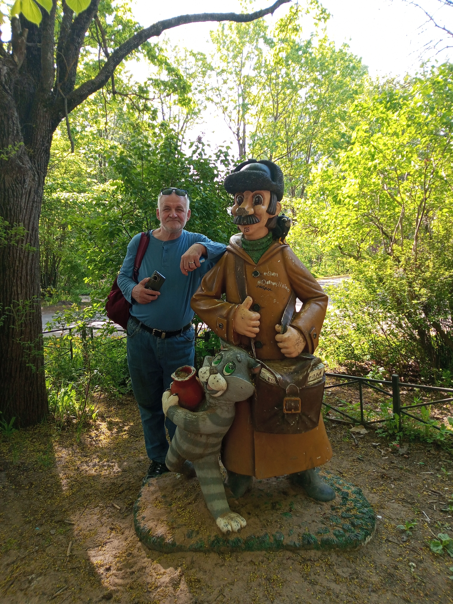 A man is posing next to a cartoon statue in what appears to be a park in Kommunar, Russia. The man, a mature adult, wears a blue long-sleeved shirt, blue jeans, and black shoes. He holds a black smartphone in his left hand and leans on the statue with his right. The statue is of a cartoonish character with a large mustache, wearing a brown coat, green turtleneck, and a black hat. A cartoon cat stands beside the character, holding a red pot. A brown messenger bag hangs from the statue's shoulder. They are on a dirt and leaf-covered area. Behind them, the scene is green with trees and foliage. A tree trunk is to the man's left and a thin metal fence is to the right of the statue. Sunglasses are resting on the man's forehead.
