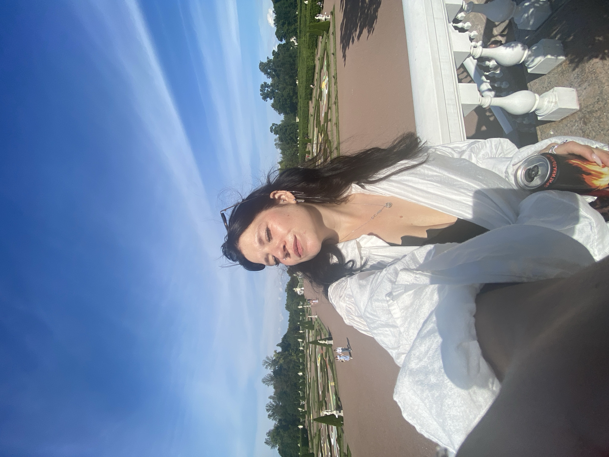 The image is a selfie taken outdoors on a sunny day, with a clear blue sky and wispy clouds. In the foreground, a woman with dark hair is holding a can of energy drink and wearing a white oversized shirt. She has a necklace and earrings. In the background, there is a meticulously maintained formal garden with manicured bushes, trees, and statues. The garden appears to be part of a larger estate or park. The architectural style of the balustrade in the foreground suggests a historical European setting. The scene is likely the formal gardens of Peterhof Palace in Lomonosov, Russia, a renowned landmark known for its elaborate landscaping and fountains.