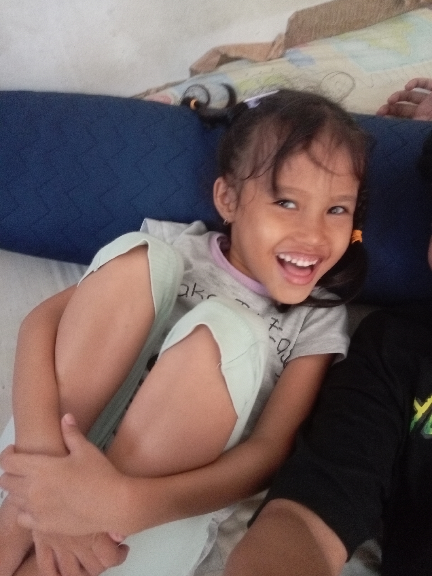 A young girl with dark hair styled in pigtails is lying down, her legs bent and crossed. She is wearing a light grey t-shirt with a design on it that is partially visible. Her head is turned towards the camera, and she has a wide, joyful smile, showing her teeth and a glimpse of her tongue. Her eyes are open and looking at the viewer, and her expression is one of happiness and amusement. She appears to be in an indoor setting, possibly a bedroom or living area, with a blue pillow or mattress behind her. The lighting suggests it might be daytime. To her right, another person's arm and shoulder, wearing a black t-shirt, are visible, suggesting they are lying next to her. The overall mood of the image is lighthearted and playful. There is some text visible on the girl's t-shirt, but it is too blurry to read.