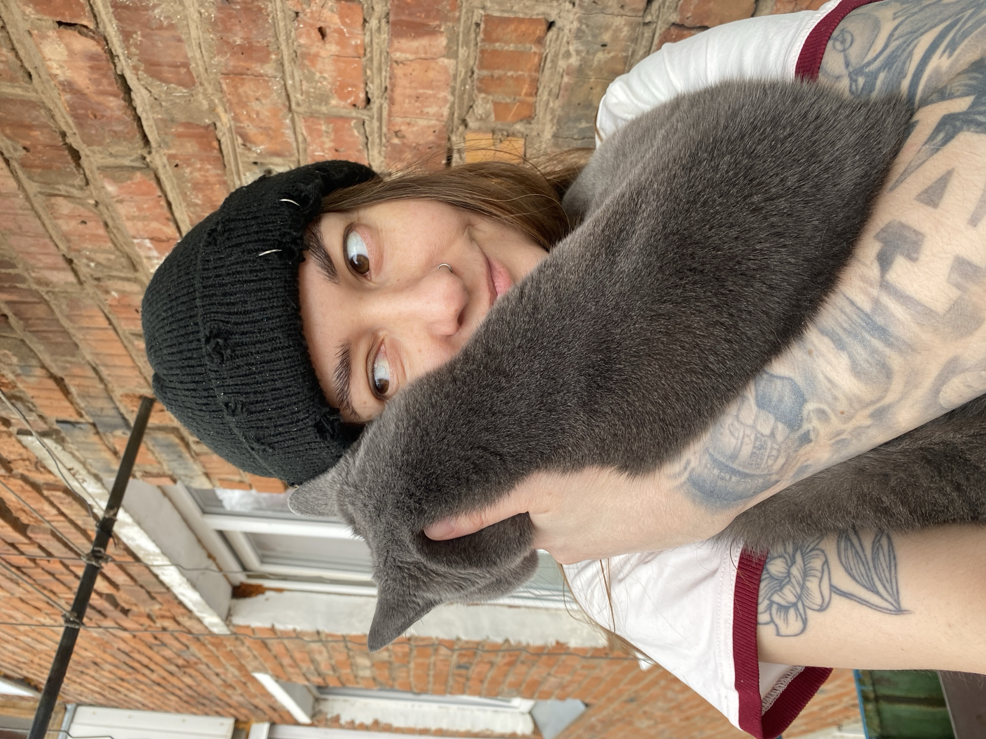 A person wearing a black beanie with metal pins and a white t-shirt with a maroon trim is holding a gray cat. The person has fair skin, brown hair, brown eyes, and a nose ring. The person's right arm is visible and has multiple tattoos. The background includes a red brick wall, a white window frame, and a metal pole with wires. It is daytime. The location is Votkinsk, Russia.
