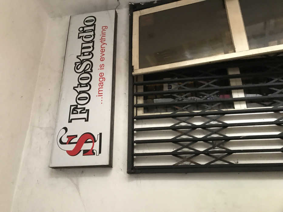 A vertical sign reads "FotoStudio...image is everything" with a stylized red "S" beneath. Adjacent to it is a window with a white frame, partially obscured by a black, expandable metal security grille. The wall appears aged and textured.