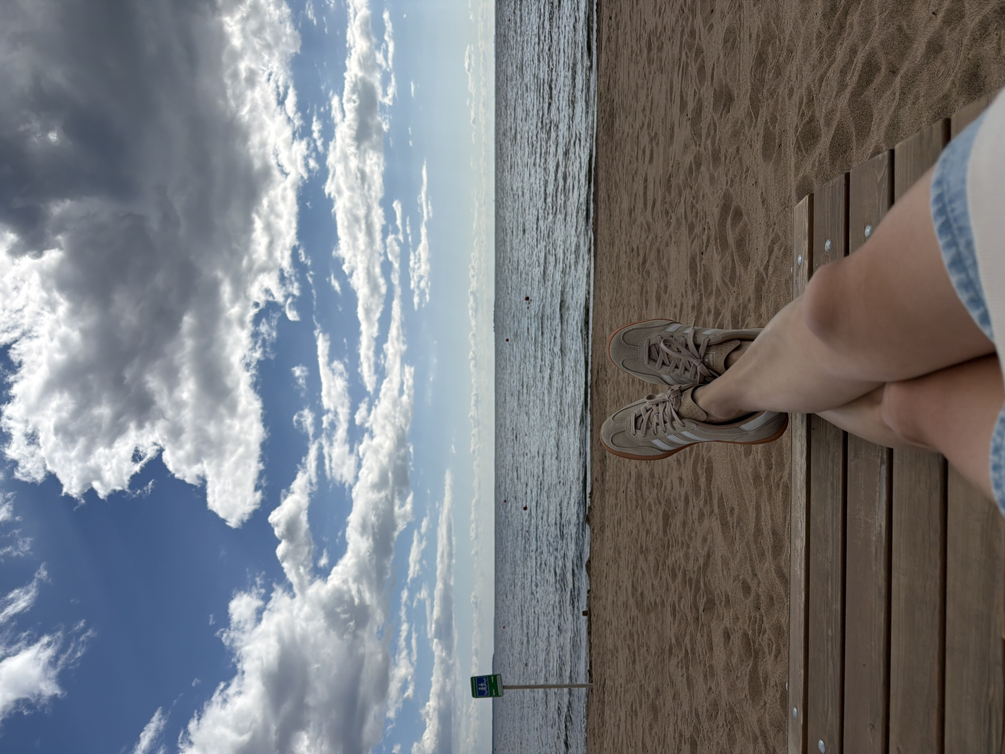 A first-person perspective shot shows a person's legs and feet resting on a wooden bench on a sandy beach. The person is wearing tan sneakers with white stripes. The beach stretches out to a calm body of water under a sky filled with dramatic, fluffy white clouds. A signpost with a green symbol is visible on the left side of the frame, near the water's edge. The scene appears to be a serene beach environment in Repino, Russia, likely during daytime given the bright sky.