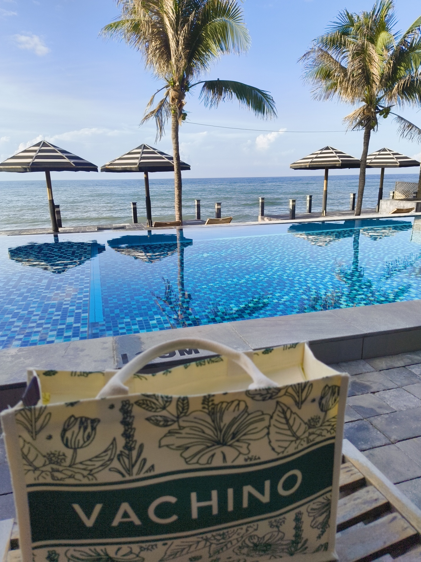 A tote bag with a floral pattern and the word "VACHINO" printed on it rests on a wooden chair in the foreground. Behind the chair, an infinity pool reflects the sky and palm trees. The pool is bordered by gray stone tiles. In the background, the ocean stretches to the horizon under a bright blue sky with scattered clouds. Three striped umbrellas are planted on the sandy shore, and two palm trees stand tall on the left and right sides of the scene, framing the view of the ocean. The overall scene depicts a serene beachside resort, likely located in Phu Thuy Ward, Vietnam, given the context.