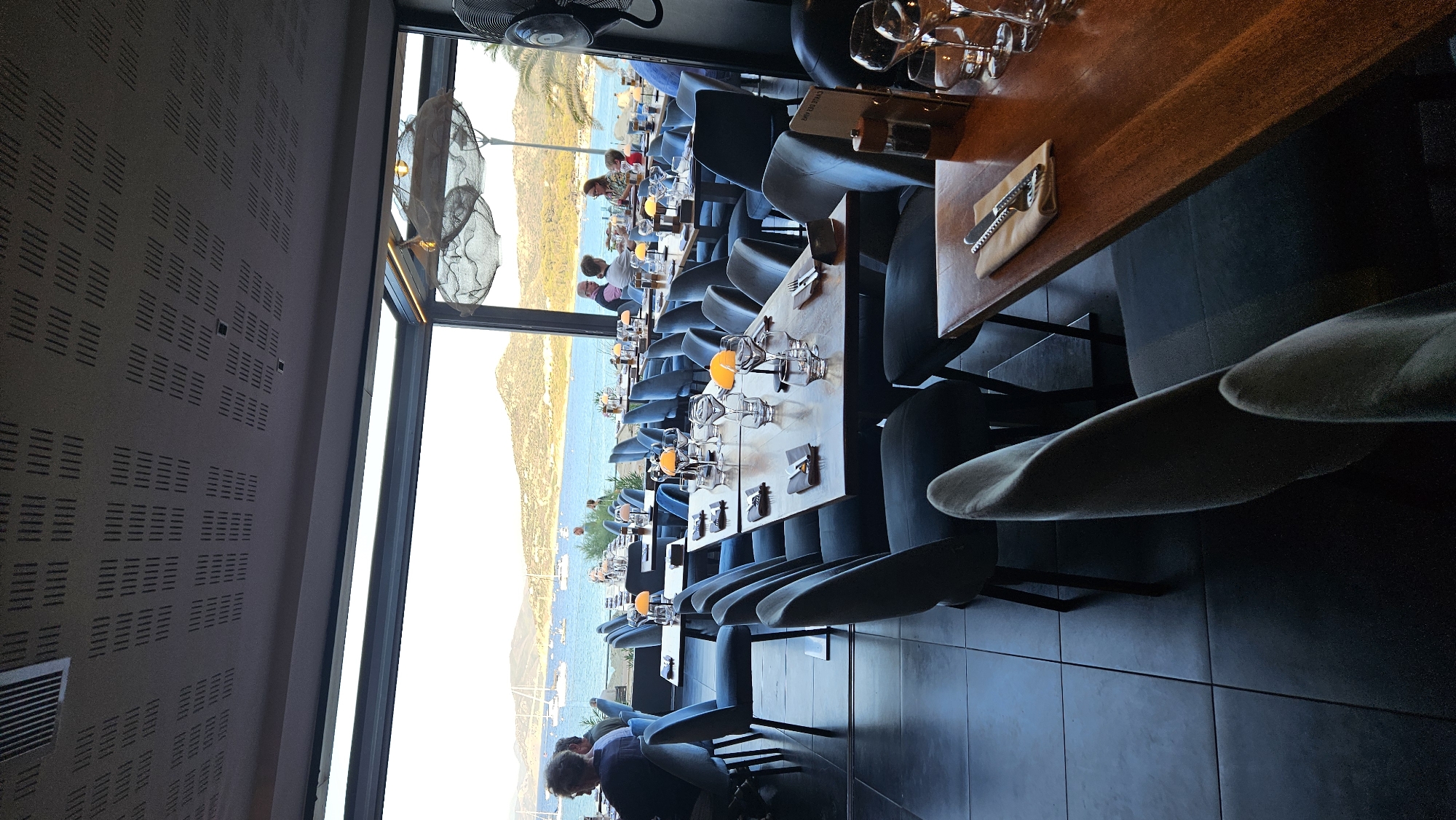 The image shows the interior of a restaurant with a view of the sea and coastline. Several tables are set with placemats, silverware, wine glasses, and small orange lamps. Blue upholstered chairs surround the tables. In the background, through a large window, sailboats are visible on the water, and a hilly, vegetated coastline is in the distance. The ceiling of the restaurant features a textured, perforated panel. The floor is tiled with large, dark square tiles. Several patrons are seated at tables, some facing the window and others looking towards the interior of the restaurant. The lighting suggests it is either late afternoon or early evening. The restaurant is located in L'Île-Rousse, France.