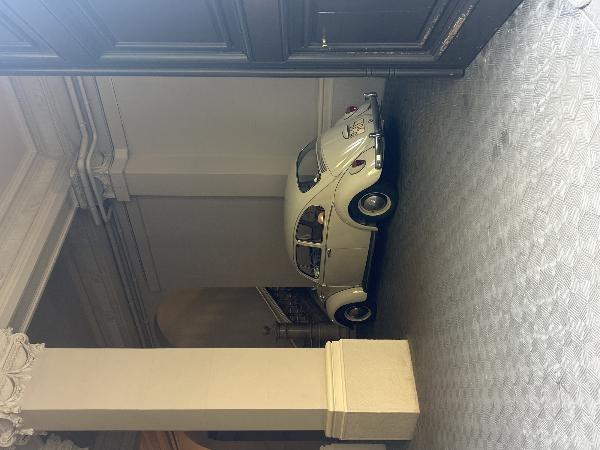 An indoor shot shows a white classic Volkswagen Beetle parked inside a building that appears to be in Barcelona, Spain. The car is neatly aligned against the wall. The license plate on the car is visible, reading "B-394 B 201". The surrounding environment includes ornate architectural elements, such as decorative columns and patterned flooring. The overall setting suggests a high-end lobby or historical building.