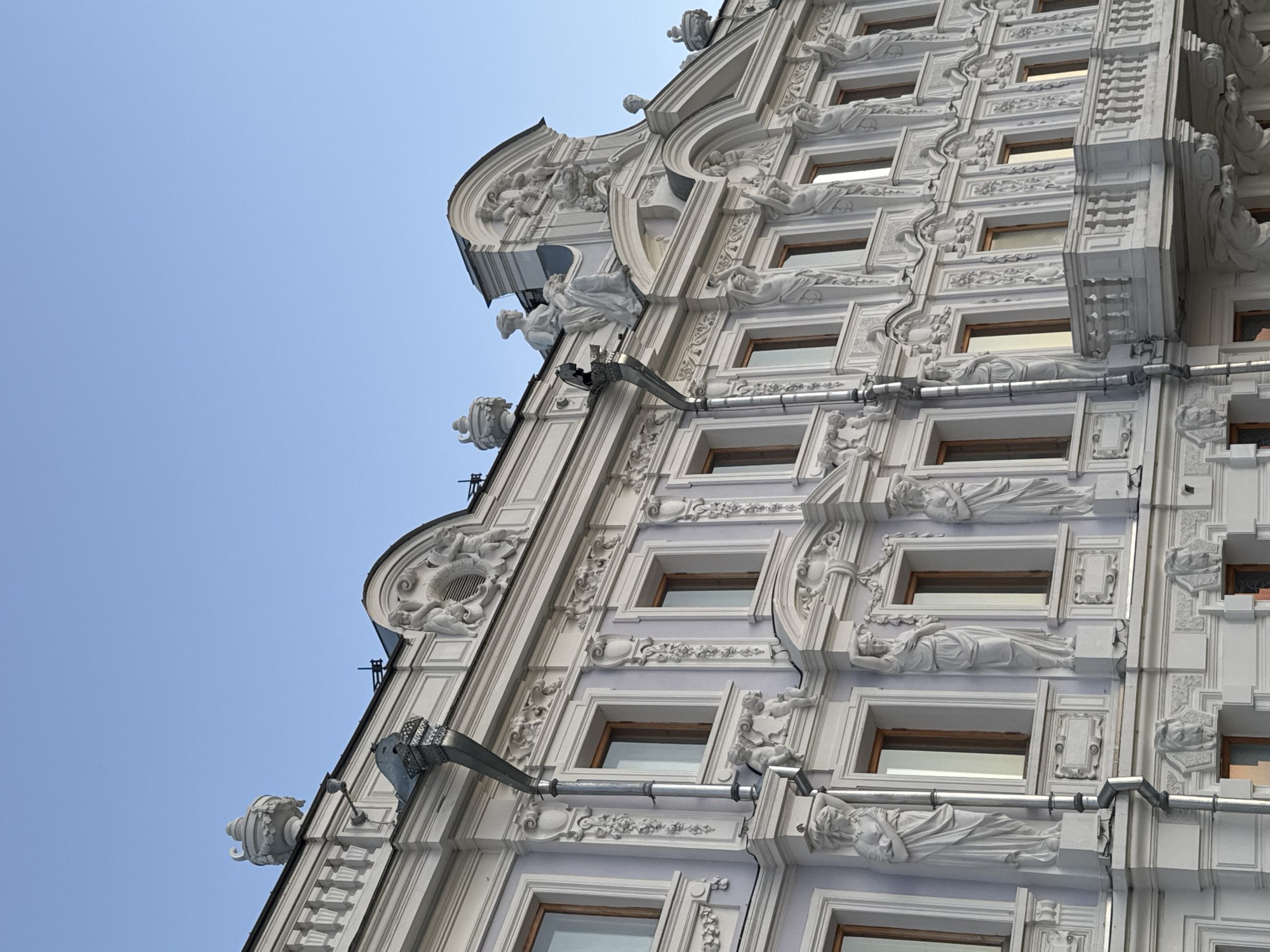 The image shows the facade of a historic building in Nizhny Novgorod, Russia. The building is adorned with intricate white stucco ornamentation, including statues of figures, decorative reliefs, and architectural elements like balconies and cornices. The sky above is clear and blue, indicating daytime. The architectural style suggests a late 19th or early 20th-century European influence, possibly Art Nouveau or Neoclassical. The building's facade is a light purple-gray color, contrasting with the white stucco. There are several windows with wooden frames visible. The overall impression is one of grandeur and artistic detail.