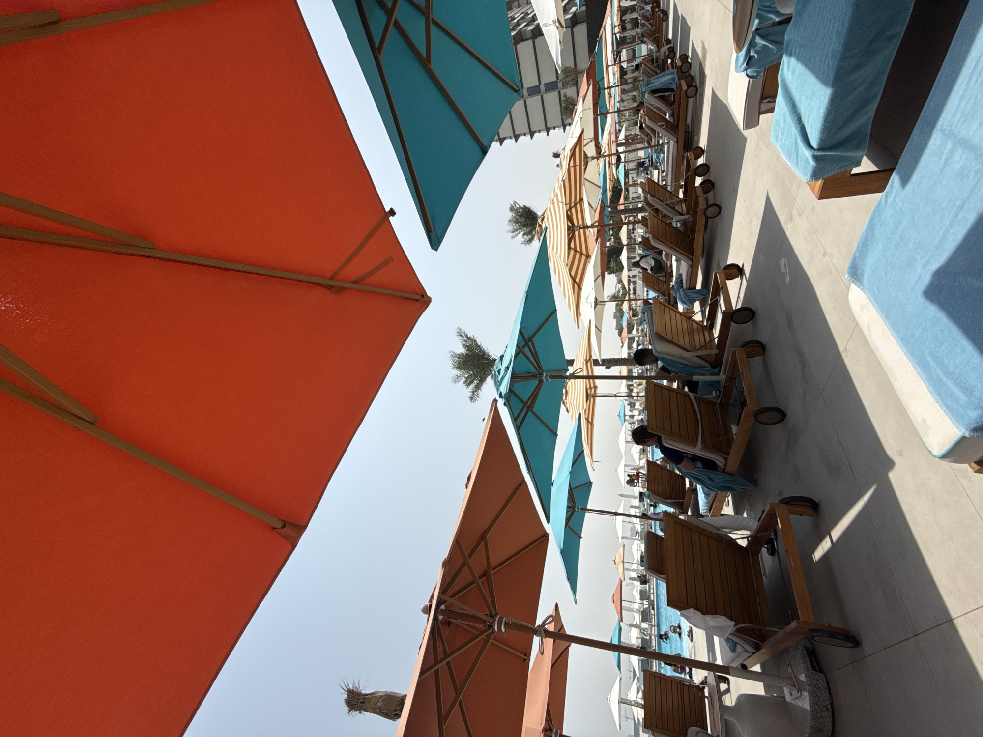 The image depicts a poolside scene at a resort. Several lounge chairs with blue towels are visible, some occupied by people. Various umbrellas, including large orange and teal ones, are providing shade. In the background, a swimming pool with people can be seen, along with more lounge chairs and umbrellas. A modern building is visible in the far background. The scene appears to be in a sunny, outdoor environment. The city is not specified but the country is the United Arab Emirates.