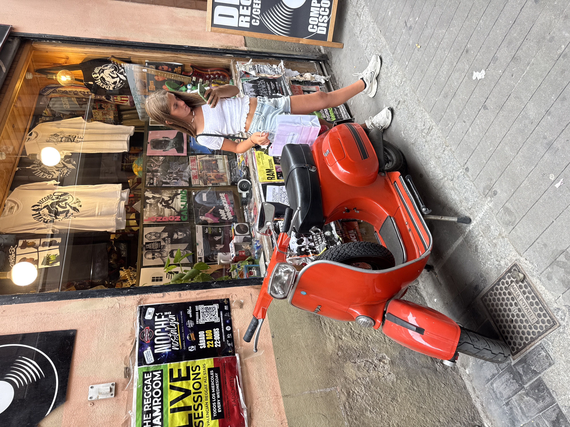 An outdoor street scene in the city of Valencia, Spain, features a young woman standing beside a bright red vintage Vespa scooter, looking at her mobile phone. She is wearing a white cropped top and light blue denim shorts, with white athletic shoes. She holds a shopping bag in her left hand. The scooter is parked on a cobblestone sidewalk in front of a shop with its window display visible. The shop window showcases an array of music records and band merchandise, including t-shirts and tote bags, with the "Devil Record" logo prominently displayed. Posters advertising events such as "Reggae Jamroom Live Sessions" and "Noche Nostalgia" are affixed to the pink-colored wall beside the shop. The street is paved with grey flagstones and cobblestones. A metal manhole cover, labeled "AGUA POTABLE," is visible on the ground near the scooter's front wheel. The overall environment suggests a lively urban area with a strong connection to music culture.