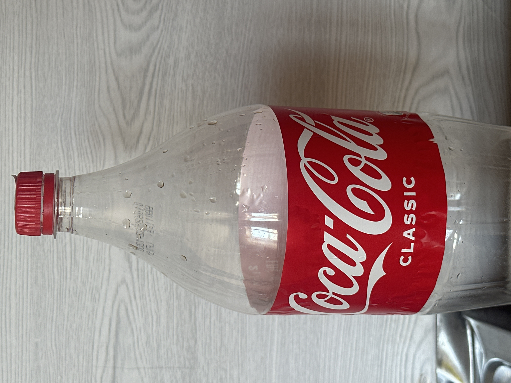 A close-up, vertical, eye-level shot shows a clear plastic bottle of Coca-Cola Classic with a red cap. The bottle is partially filled with a pinkish liquid and has condensation on its exterior. The Coca-Cola logo is prominently displayed in white script on a red label. Behind the bottle, a light-colored wooden-paneled wall is visible, characterized by horizontal lines. In the lower-left corner of the frame, a portion of a metallic object, possibly a sink or basin, is present. The image does not contain any people, animals, or indications of a specific location within the city of Gyumri, Armenia, or any other identifiable geographical setting.