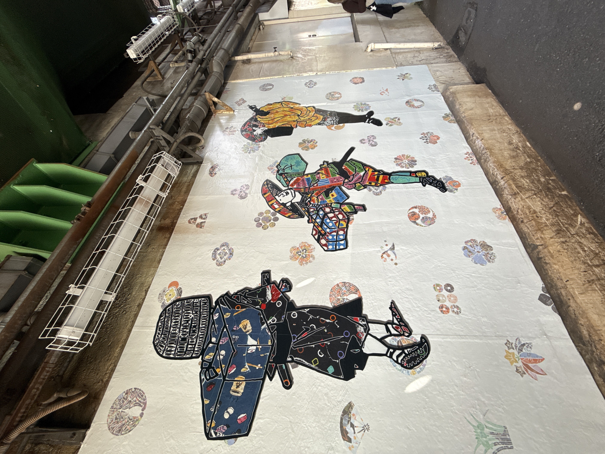 The image captures a floor in Shibuya, Japan, displaying three intricate figures or sculptures laid flat. The figures appear to be stylized human forms adorned with geometric patterns, cultural symbols, and vivid colors. They are arranged linearly on a light-colored surface patterned with smaller decorative motifs. The environment is industrial, possibly an interior space with metallic structures, pipes, and lighting fixtures visible on the left side of the image. The composition is shot from a skewed perspective, looking down at an angle towards the floor, adding a sense of depth and emphasizing the unusual art installation in this urban setting. A wooden wall is visible on the right.