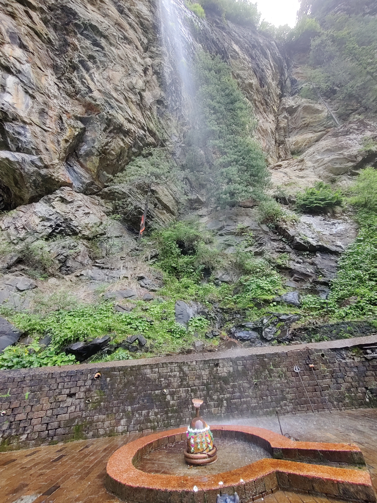 A vertical, eye-level shot captures a naturalistic scene of a waterfall cascading down a sheer rock face in Majhach, India. In the foreground, a circular stone structure, possibly a shrine or fountain, contains a Shiva lingam adorned with garlands and a snake motif at its base. The structure is surrounded by water, and water sprays upwards, creating a misty effect. Behind this, a brick wall with embedded decorative elements runs horizontally across the frame. The background is dominated by a rugged, textured cliff face, covered in patches of green vegetation and trees, with the waterfall a thin, white stream originating from the top. The lighting is overcast, suggesting a cloudy day.