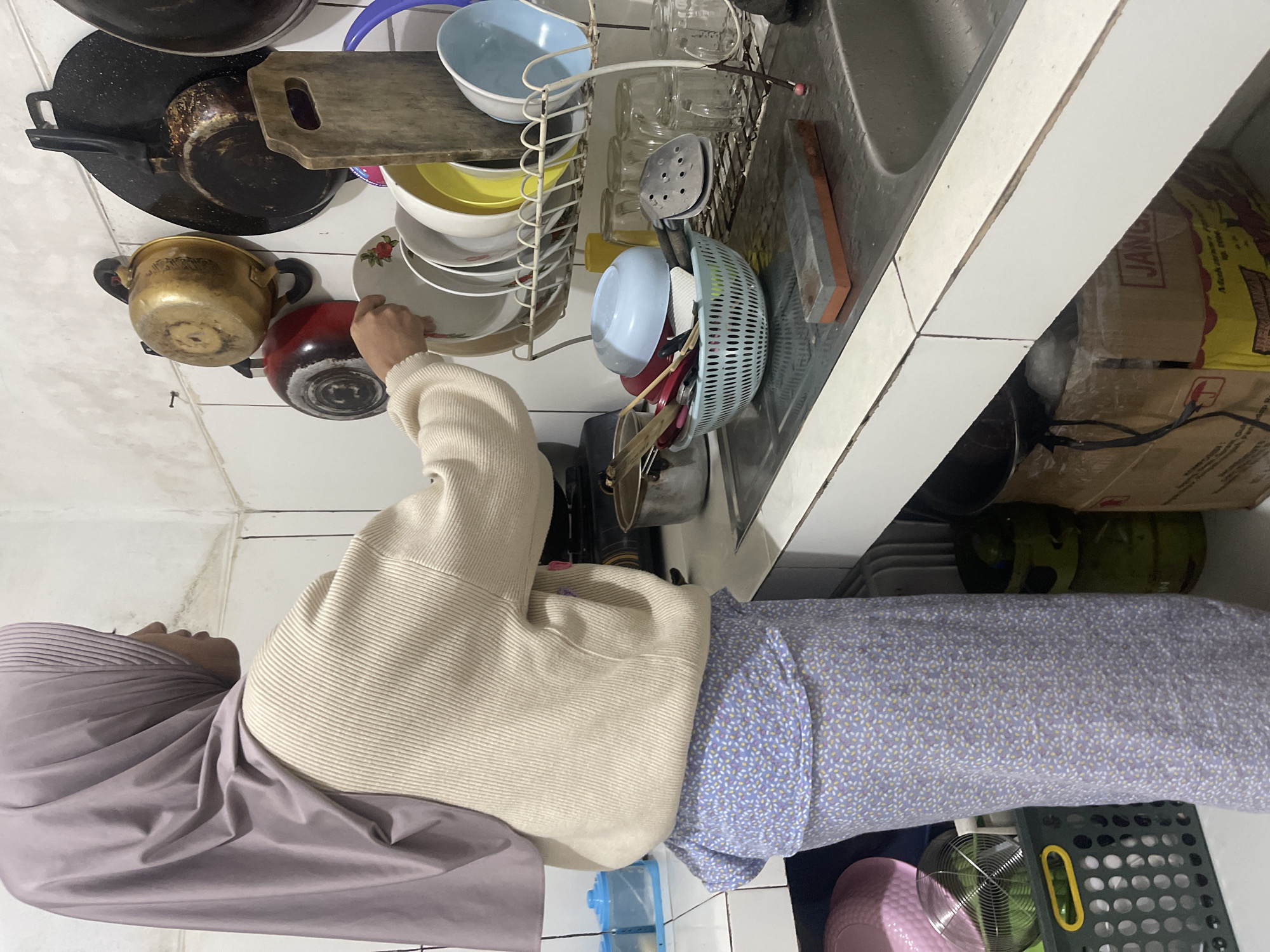 A woman wearing a light purple hijab and a cream-colored knitted sweater over a light blue floral patterned skirt is standing in a kitchen in Gunung Sindur, Indonesia. She is reaching towards a drying rack filled with white and yellow plates and blue and white bowls. To the left of the drying rack, there is a white sink with a silver faucet. On the counter next to the sink, there is a sharpening stone. Above the drying rack, several pots and pans are hanging on hooks mounted on the wall. The kitchen appears to be functional with various kitchen items visible.