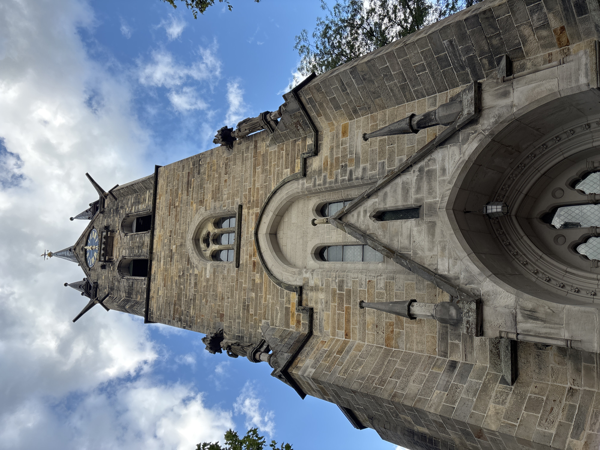 A low-angle, wide shot captures the stone tower of a gothic-style church against a bright blue sky with scattered white clouds. The tower, constructed from light brown and grey stone blocks, features a central clock face with Roman numerals and ornate hands. Flanking the clock are two arched windows, and above it, a steepled roof with decorative elements and a weather vane. The tower is adorned with gargoyles, statues, and pointed spires. A large arched entranceway with decorative stonework is visible at the base of the tower, with a lantern hanging from the arch. Trees can be seen on the right side of the frame. The scene is static and depicts the exterior architecture of the church.