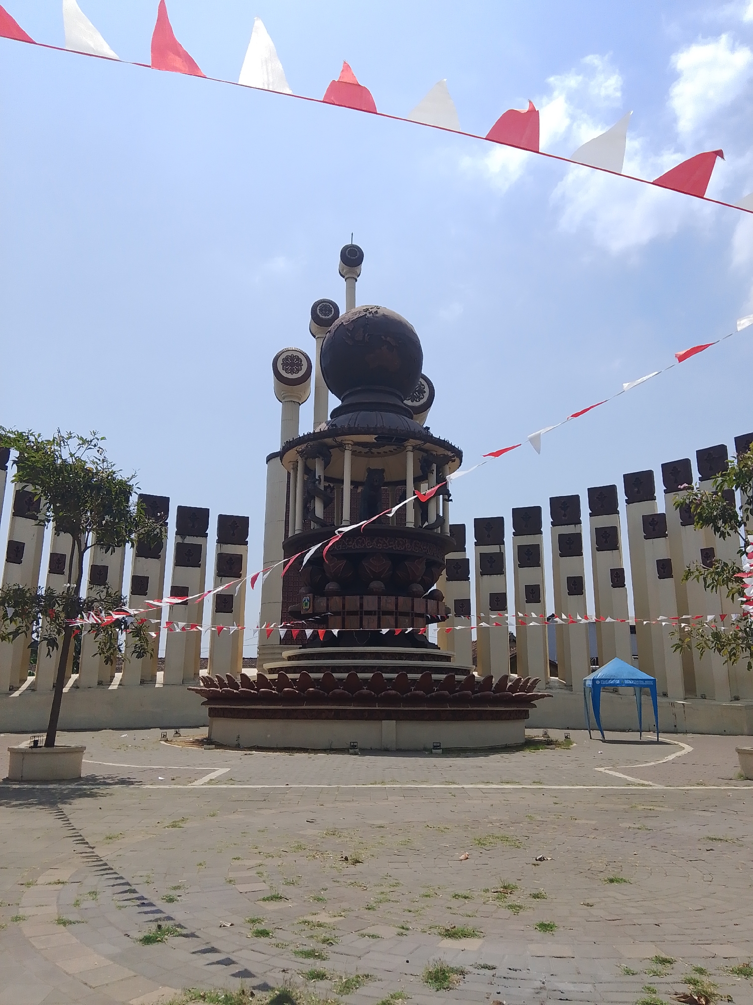 The image captures a monument in Gedangan, Indonesia, under a clear sky with scattered clouds. Red and white triangular flags are strung across the scene, creating a festive atmosphere. The monument itself is a large, ornate structure with multiple tiers and decorative elements. At its center is a spherical object, possibly a globe, atop a dark, intricately carved base. Surrounding the central monument is a semi-circular arrangement of tall, slender pillars, each topped with a dark, square cap. These pillars are interspersed with what appear to be smaller, decorative rectangular blocks. Trees with green foliage are visible on the left side of the image, and a small blue tent is set up in the background to the right. The ground is paved with square tiles, and patches of grass are growing through the cracks. Shadows from the monument and flags are cast on the paved area, indicating sunlight. There are no people visible in the image.
