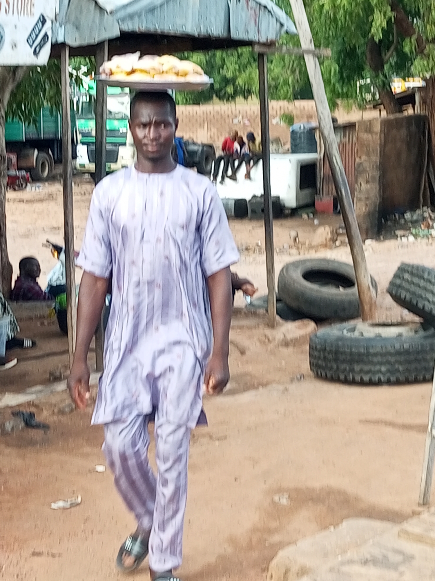 The image depicts an outdoor scene in an unpaved area in the city of Unknown, Unknown, likely an informal market or residential street. The ground consists of dry, reddish-brown earth, uneven in texture and showing signs of foot traffic, with scattered small pieces of white litter.
In the foreground, an adult male with a dark complexion walks towards the viewer, positioned slightly to the right of the central axis. He wears a light lavender-colored traditional tunic (buba) with vertical stripes and matching trousers (sokoto), and dark open-toed sandals. He carries a flat, silver-colored tray balanced on his head, laden with numerous light-colored, possibly baked goods or bread rolls. His gaze is directed forward, and his brow is slightly furrowed. His arms hang relaxed at his sides.
To the left of the main subject, a rustic structure is partially visible, featuring a dark wooden post supporting a corrugated metal roof. A white sign with the word "STORE" and some illegible text is affixed to this post. Further in the background, beneath this shelter, parts of a large, dark green truck are visible, including a rear wheel and chassis. Two individuals are partially visible sitting in the shade under the roof structure in this area.
On the right side of the mid-ground, a thick, light-colored wooden pole or beam extends diagonally from the upper right toward the lower center, partially obscured by the main subject. Several used vehicle tires are scattered on the ground; one lies flat on its side, and two others are positioned closer to the foreground. Behind these tires, a low, rectangular white structure is visible, followed by a dark blue water tank supported on a stand. To the right of the water tank, a rudimentary building with light-brown, possibly mud-brick walls and a section of corrugated metal siding is present. Further back on the left, a group of approximately three to four individuals are seated on a low platform or ledge under a small, simple shelter.
The background is dominated by dense green foliage from trees, indicating a natural environment beyond the immediate structures. The scene is illuminated by natural daylight.
