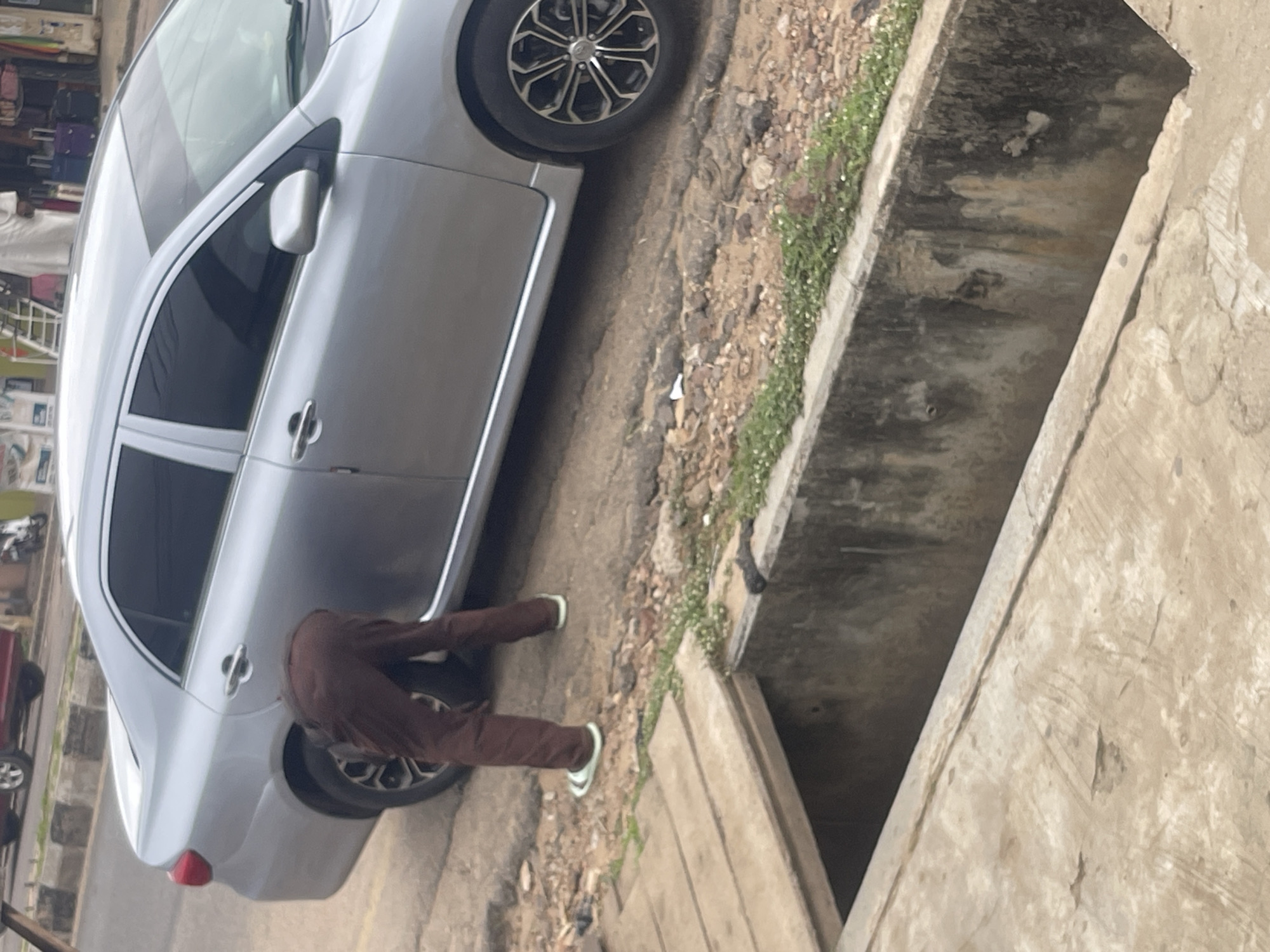 A person is seen bending over next to a silver sedan parked on a street in an urban environment. The person is wearing brown pants and light-colored sandals. They appear to be attending to the rear tire of the car. In the foreground, there is a concrete drainage ditch. The background shows a sidewalk and some buildings. The lighting suggests it is daytime.