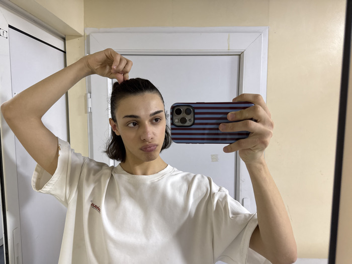 A person is depicted from the chest up, taking a mirror selfie indoors. The individual has short, dark hair, which is pulled back and held at the crown by their right hand. They wear a light-colored, possibly white, loose-fitting t-shirt with a mirrored red logo or text on the chest, and the right sleeve exhibits minor tearing. Their left hand holds a dark smartphone, featuring a triple camera array and a case with horizontal burgundy and light blue stripes. The individual's gaze is directed slightly to their left, with a neutral expression. The reflected background includes a white door or cabinet with a dark vertical frame to the left of the subject, and a plain, light beige wall to the right. No specific identifiable markers within the image indicate the location as Yerevan, Armenia.