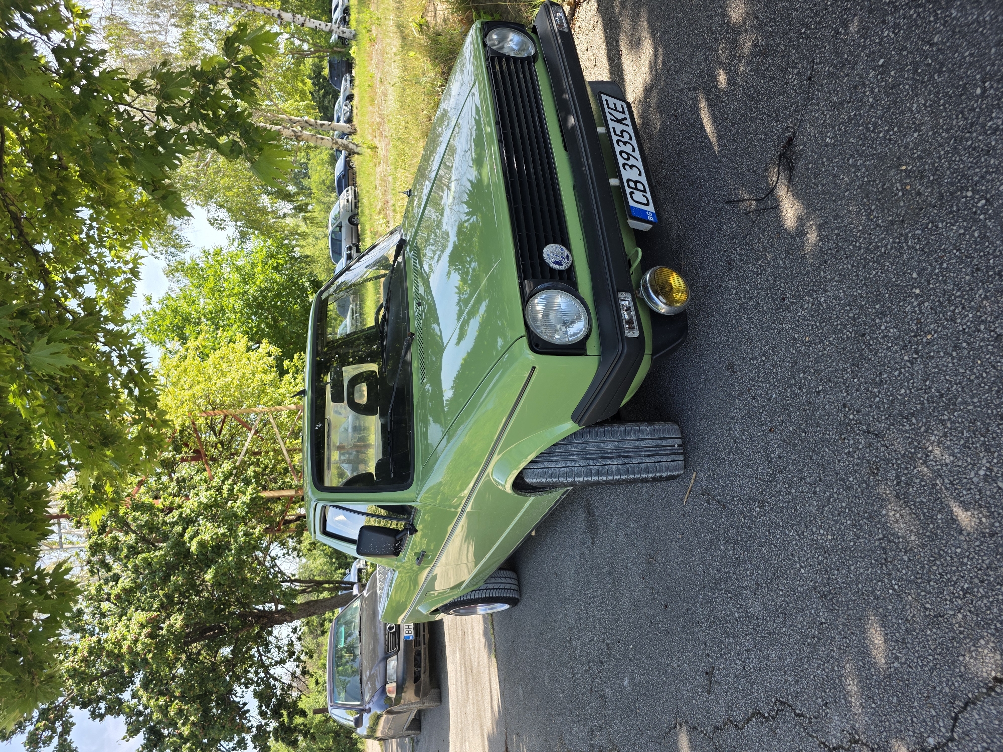 The image shows a bright green, vintage pickup truck parked on an asphalt road. The truck has a black grille, round headlights, and a yellow fog light mounted on the front bumper. The license plate reads "CB 3935 KE" with a blue and yellow BG indicator. A dark-colored sedan is parked behind the truck. The scene is set outdoors, with trees and grass in the background. The sun is shining, casting shadows on the road. The location is identified as Akatsievo, Bulgaria.