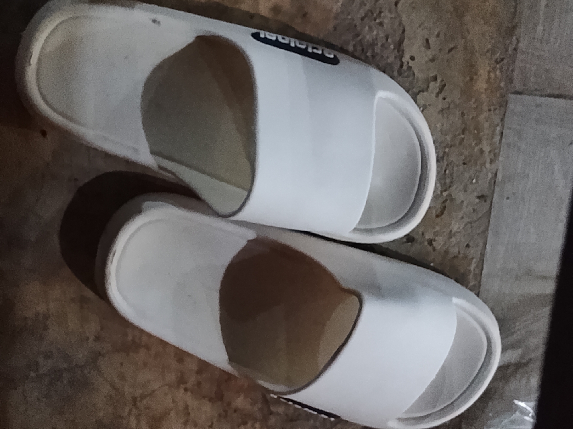 The image shows a pair of white slide sandals on a textured floor. The sandals are plain white with a black band across the top. One sandal has the word "Adidas" printed on the black band. The floor appears to be made of concrete or stone and has an uneven, mottled texture. The lighting is dim, casting shadows and highlighting the texture of the floor and the glossiness of the sandals. No people or actions are visible in the image. The location is described as Dayahlhoksukon Barat, Indonesia, but there are no visual cues in the image to confirm this specific location.
