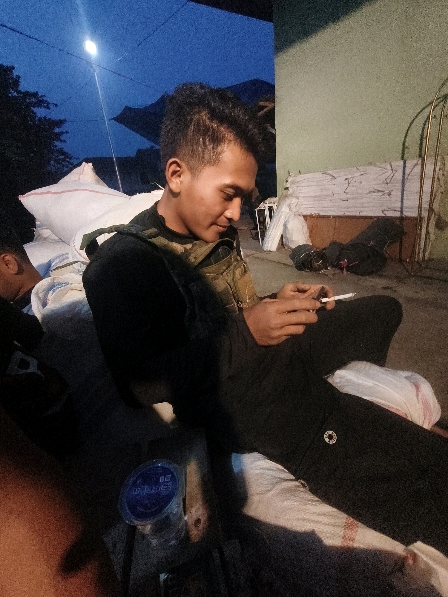 The image depicts a young man sitting outdoors at dusk, holding a cigarette. He is wearing a black long-sleeved shirt and dark pants, with a tactical vest over his shirt. The man is looking down at the cigarette in his right hand, which is lit and held between his index and middle fingers. His left hand is partially visible, also near the cigarette. He is seated on what appears to be large white sacks, possibly of rice or other goods.
In the background, to the man's left, another person is partially visible, their head and shoulder resting on a similar sack. Behind the seated man, there are more sacks and coiled dark material, possibly tarps or ropes. A pale green wall is visible to the right of the frame, with some metal framing or structure against it. The sky is a deep blue, indicating twilight, and a single bright light source is visible high up in the sky, casting a distinct shadow. In the lower left of the image, a clear plastic cup with a blue lid and some writing is visible.
The scene appears to be an outdoor storage or staging area, possibly near a building or warehouse. The lighting suggests it is evening. The context of the location being "Sungai Penuh, Indonesia" places this scene in a specific geographic and cultural setting, though the visual elements themselves do not provide explicit markers of this location beyond being a general outdoor area. The presence of sacks of goods and the overall atmosphere suggest a place of commerce or work, possibly in a rural or semi-urban area.