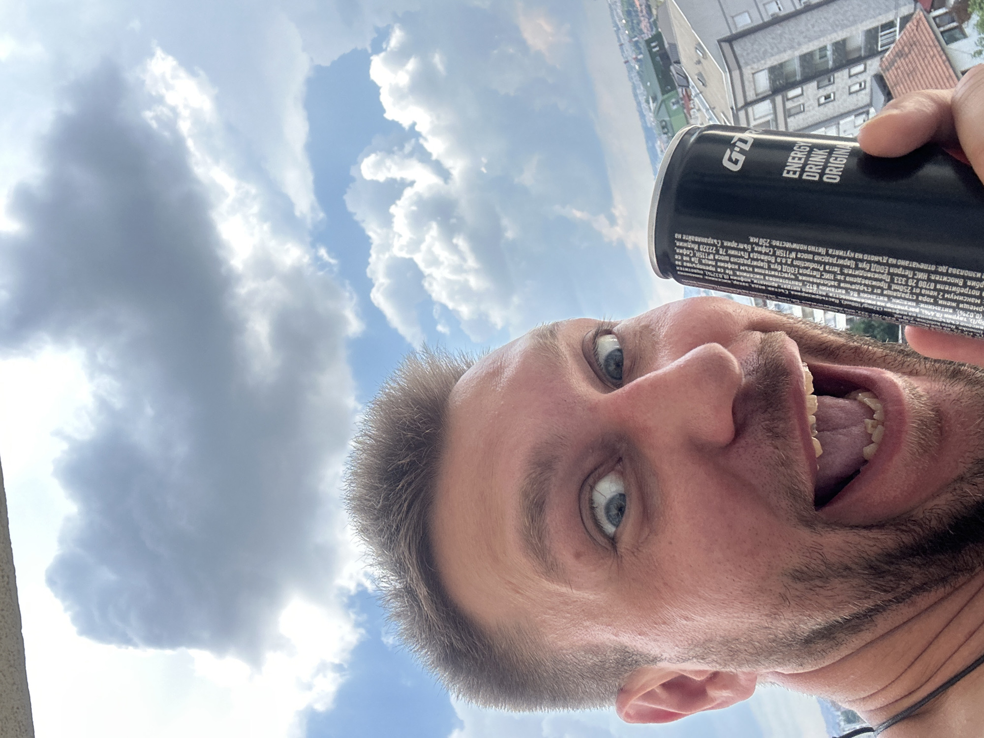 A close-up, low-angle selfie of a man holding a black can of "GDRO ENERGY DRINK ORIGINAL" in front of a cloudy sky. The man has short, spiky brown hair, blue eyes, and a beard. He is smiling widely with his mouth open, revealing his teeth and tongue. The background shows a cityscape with buildings and a clear sky filled with cumulus clouds. The location is the Zvezdara Urban Municipality in Serbia, as evidenced by the surrounding urban landscape.