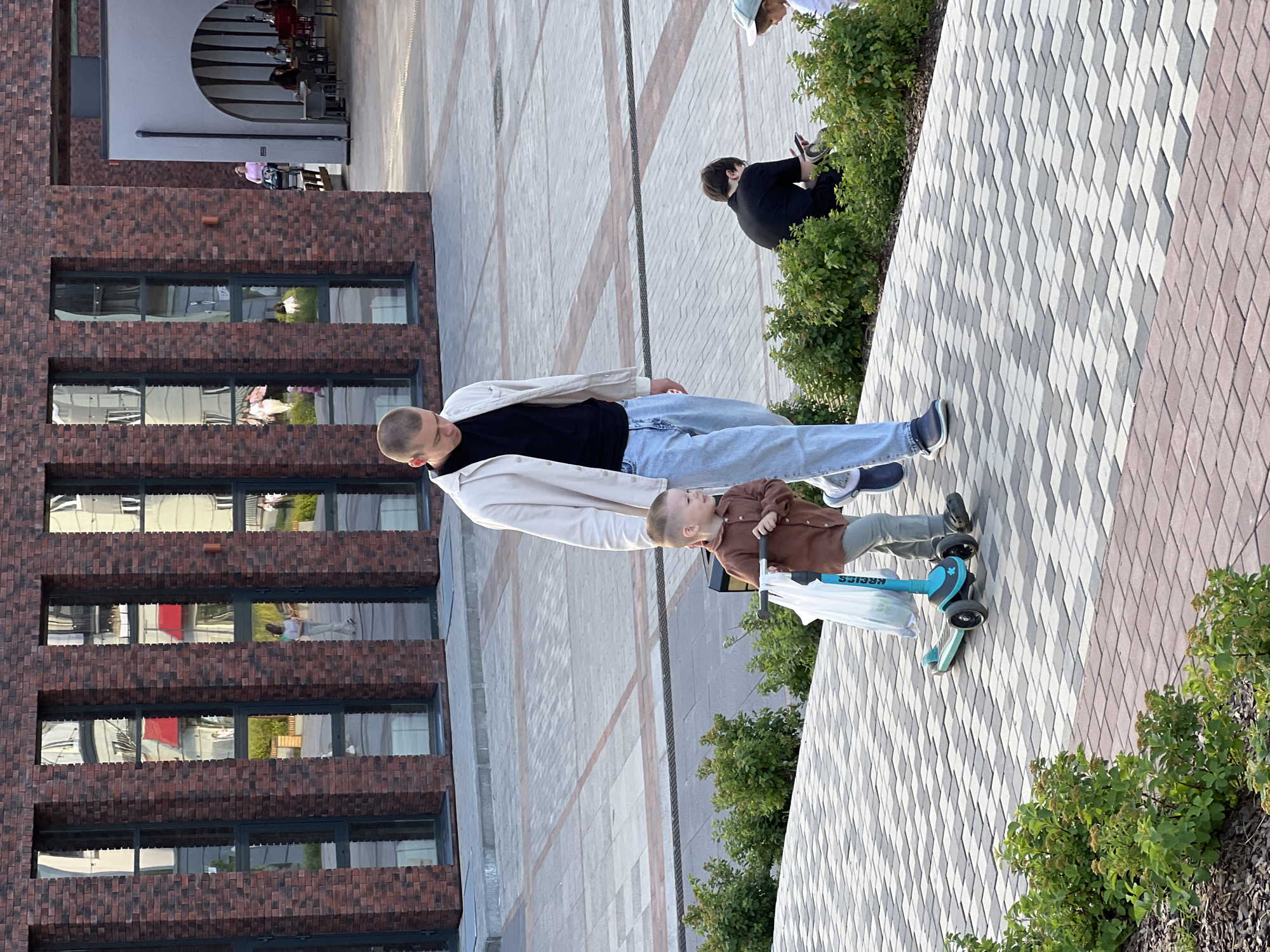 The image shows an outdoor scene with a focus on two people and a scooter. A man wearing a light-colored jacket over a black shirt and light blue jeans stands on a patterned walkway. Next to him, a young child in a brown shirt and light green pants rides a blue scooter. The child is looking up and to the right, with a white plastic bag hanging from the scooter's handlebars.
Behind the people is a modern brick building with multiple windows. Reflections in the windows show parts of the building's interior, including glimpses of people and red elements. To the right of the walkway, there is lush green foliage. The walkway itself is paved with a pattern of light and dark gray diamond-shaped tiles, bordered by darker red tiles.
Further in the background, another person in a black outfit is visible sitting amongst some bushes and looking at a phone. In the distance, through an arched opening in a building, several people are seated at tables, suggesting a cafe or restaurant area.
The lighting suggests it is daytime, with shadows visible on the ground. The overall atmosphere appears casual and relaxed, with the man and child enjoying an outdoor activity. The image is oriented vertically. There is some visible text on the scooter that appears to read "KREISS".