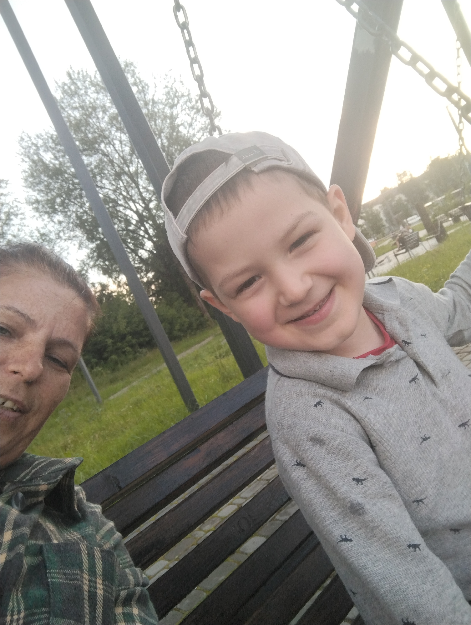 A woman and a young boy are seated on a wooden bench, likely in a park in Kingisepp, Russia, given the context. The woman is on the left, facing the camera and smiling. She has her hair pulled back and is wearing a green and black plaid shirt. The boy is on the right, also smiling broadly and looking at the camera. He is wearing a grey baseball cap and a grey long-sleeved shirt with a small pattern of dark figures on it. They are positioned close together on the bench. Behind them, trees and grass are visible, suggesting an outdoor setting. Chains of a swing set are also visible in the upper portion of the image. The lighting suggests it is daytime, possibly late afternoon or early evening due to the warm light.